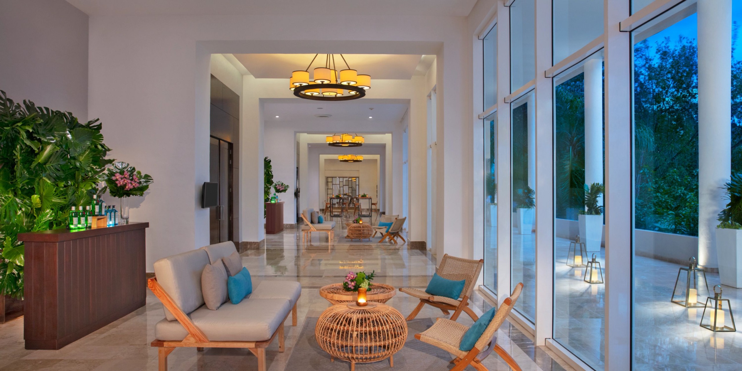 The lobby at Dreams Jade Riviera Cancun features modern wicker furniture, a sleek wooden bar, and expansive windows offering views of lush greenery. Indoor plants enhance the natural ambiance, while hanging lights beautifully illuminate the area.