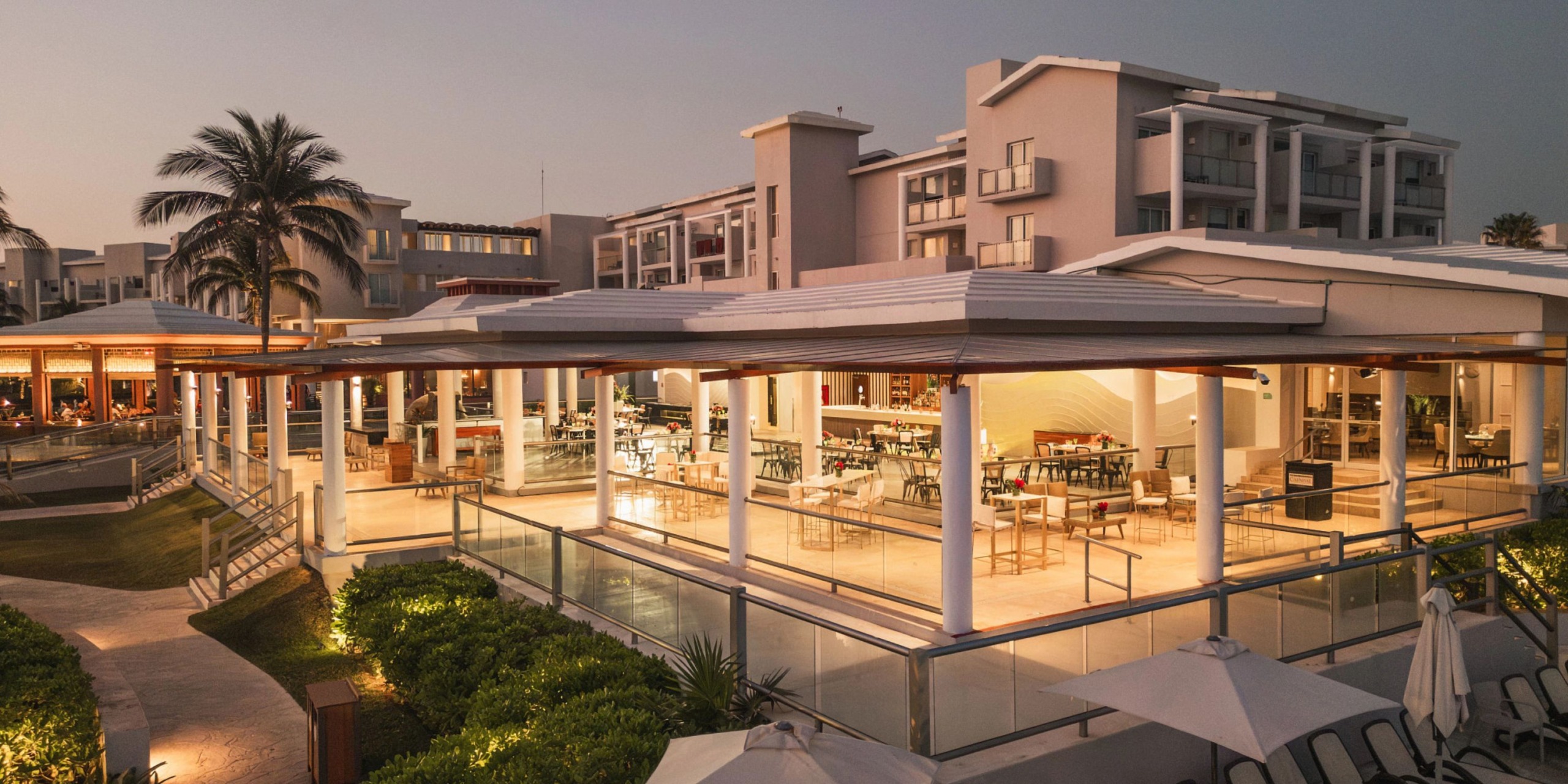 The Dreams Jade Riviera Cancun resort, featuring a lit outdoor dining area nestled among palm trees and lush greenery, is beautifully illuminated at dusk.
