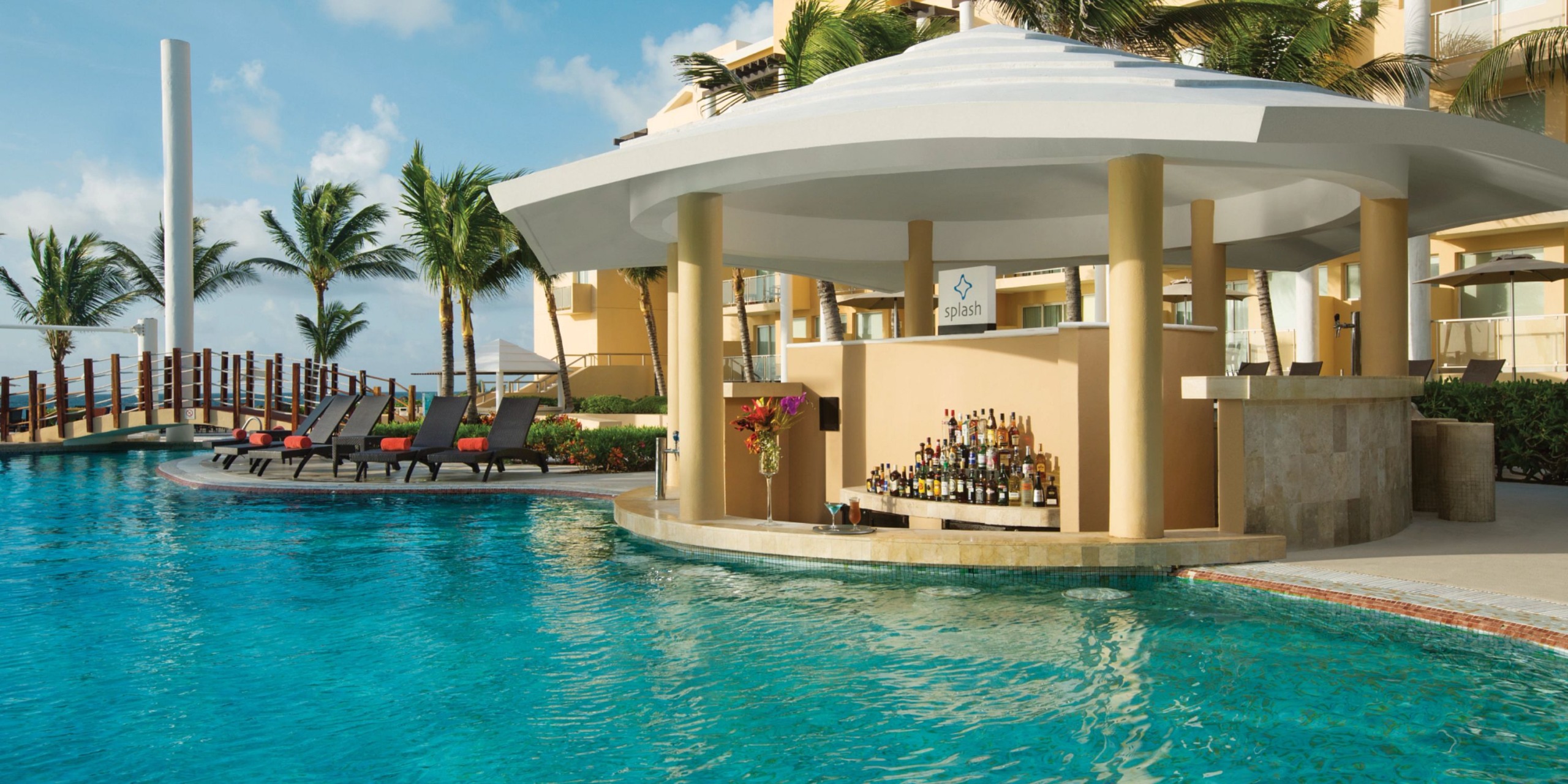 At Dreams Jade Riviera Cancun, enjoy a poolside bar offering a variety of drinks, surrounded by lounge chairs and palm trees under a clear blue sky.