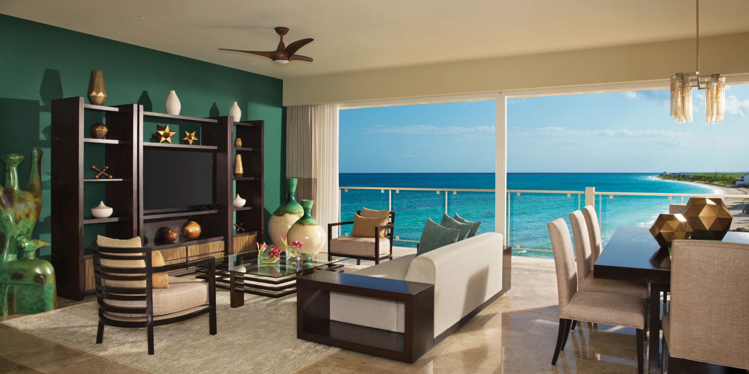 Experience the stunning modern living and dining area at Dreams Jade Riviera Cancun, complete with an ocean view, a teal accent wall, elegant wooden furniture, expansive windows, and tasteful decorative elements.