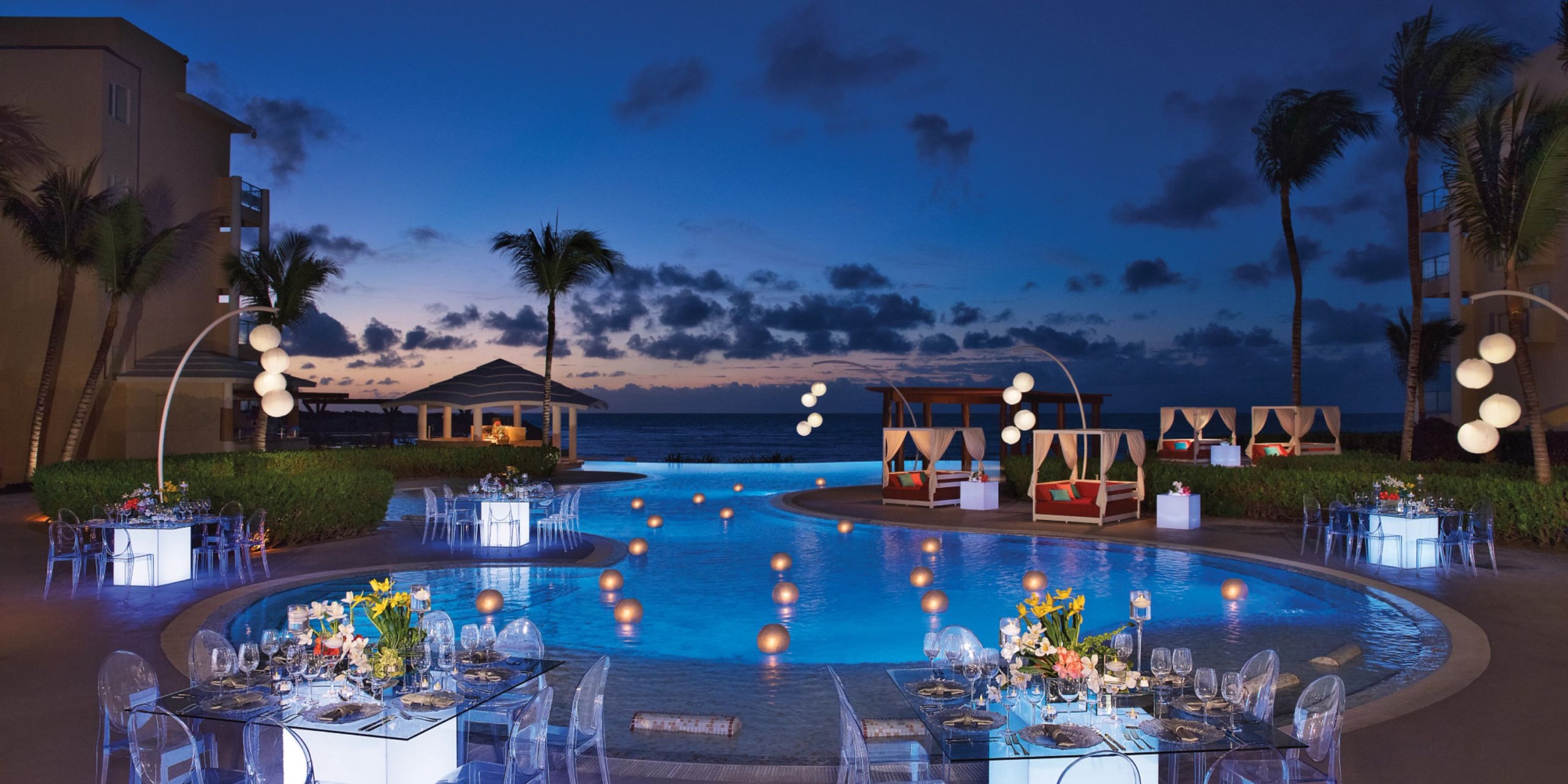 Experience an enchanting evening at Dreams Jade Riviera Cancun, featuring beautifully decorated tables by the poolside, glowing orbs floating on the water, elegant cabanas, and majestic palm trees silhouetted against a twilight sky.