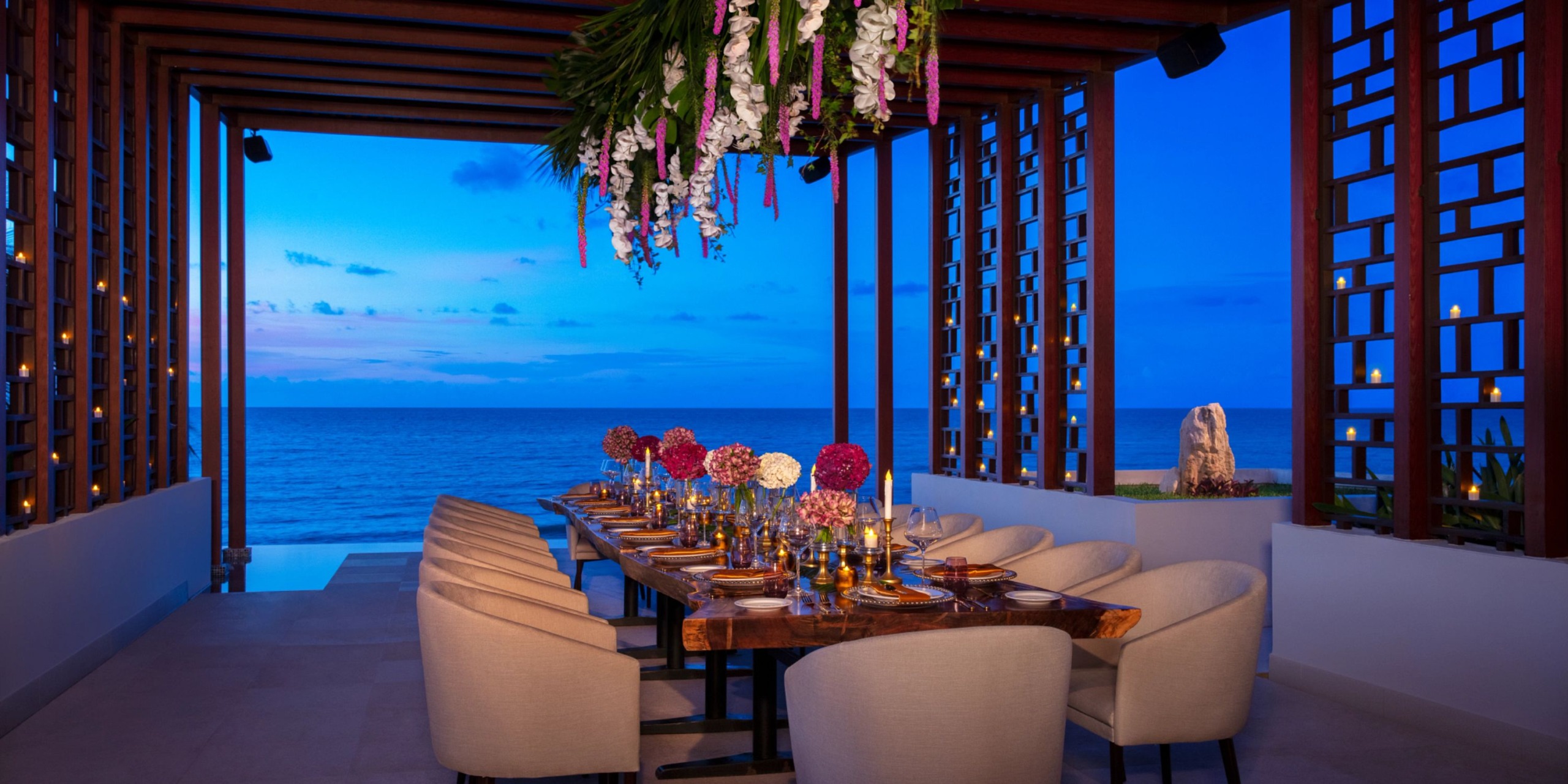 Experience an elegant dining area at Dreams Jade Riviera Cancun, featuring a long table beautifully set for dinner. Enjoy breathtaking views of the ocean at dusk while hanging flowers add a decorative touch under the open wooden roof.