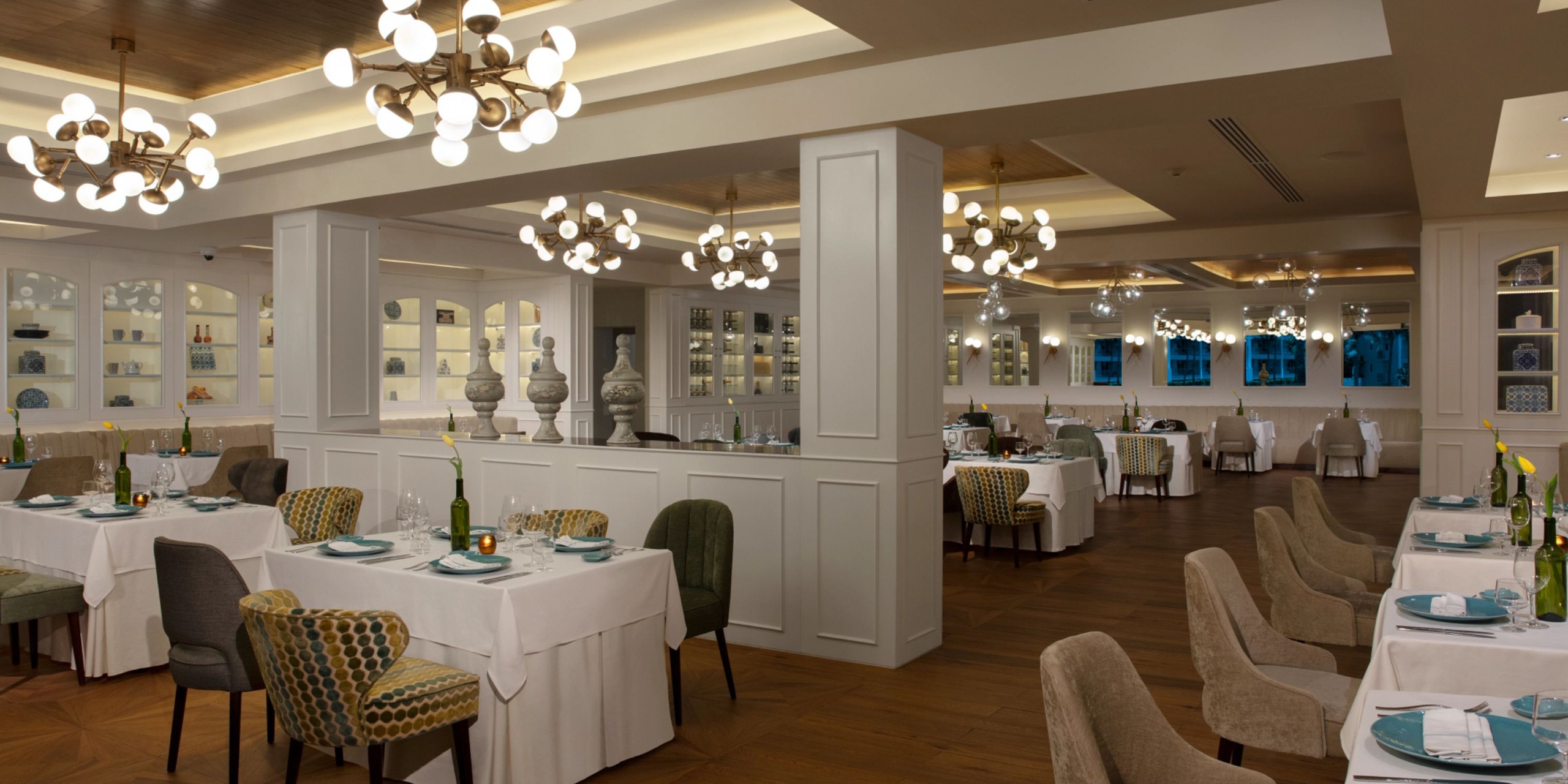 Dreams Jade Riviera Cancun offers a stylish, well-lit dining experience with white tablecloths, elegant chairs, and modern chandeliers. The tables are set for dining against a backdrop of decorative shelves.