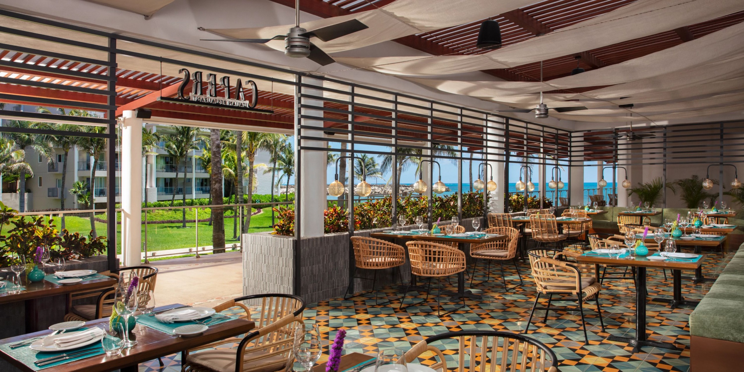 Dreams Jade Riviera Cancun features an open-air restaurant with wooden tables and chairs, decorative floor tiles, ceiling fans, and a stunning ocean view. The surrounding greenery enhances the tropical atmosphere.
