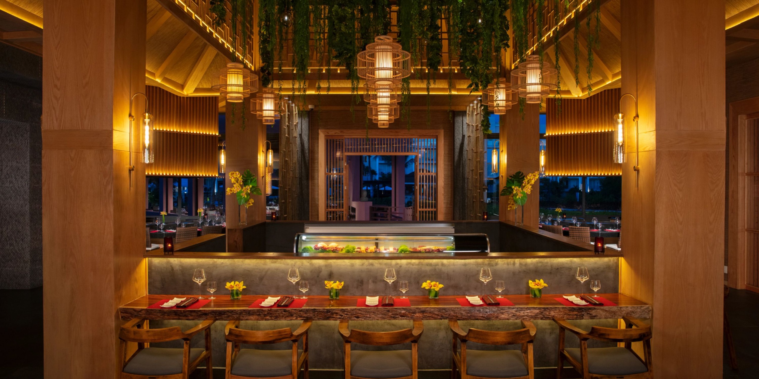 The Dreams Jade Riviera Cancun features an elegant restaurant interior adorned with wooden furniture, a central sushi bar, hanging lanterns, and decorative plants. Wine glasses and yellow flowers gracefully line the counter.