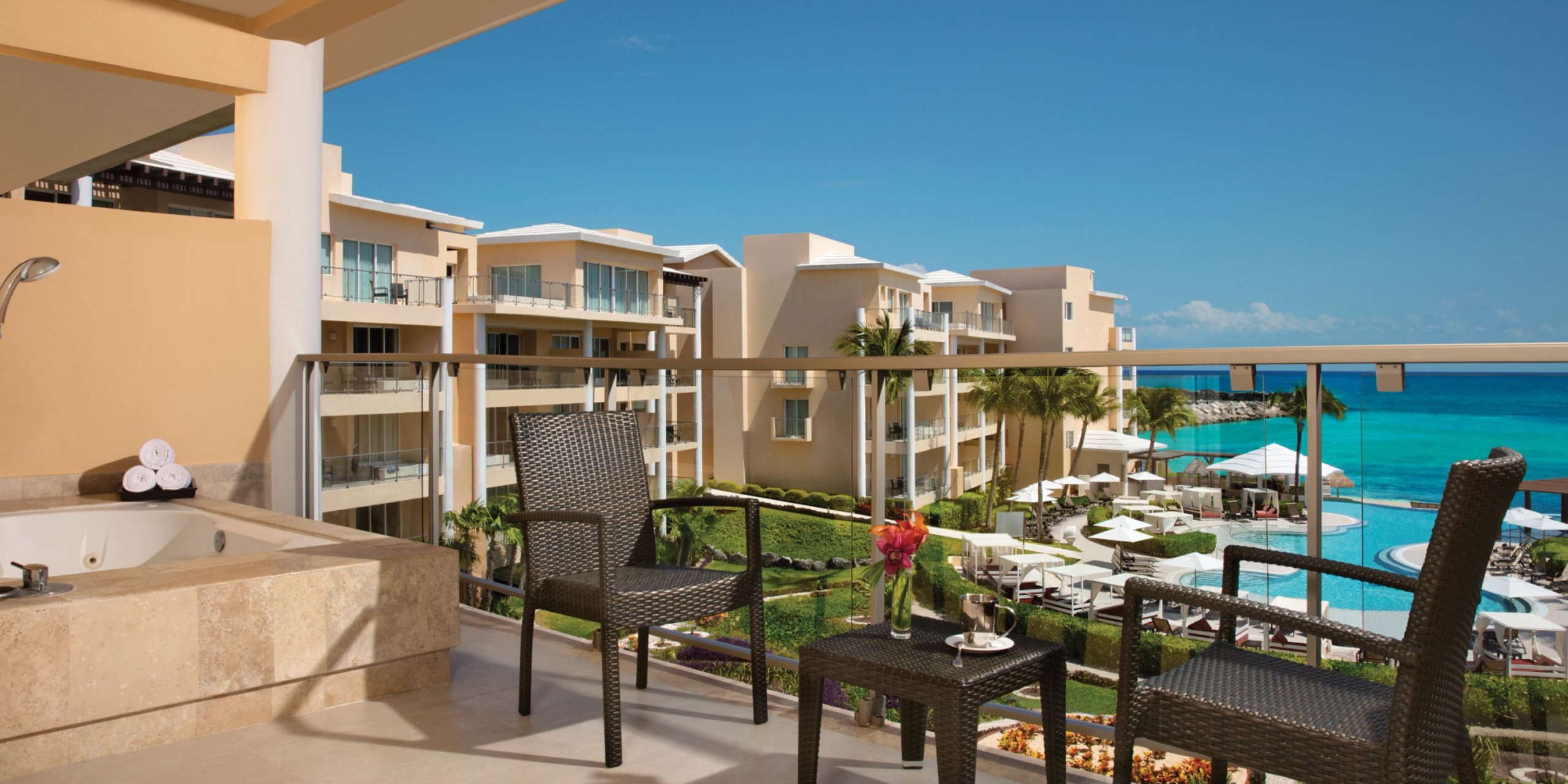 A balcony view at Dreams Jade Riviera Cancun features wicker chairs and a table, providing a stunning overlook of the pool area and ocean.