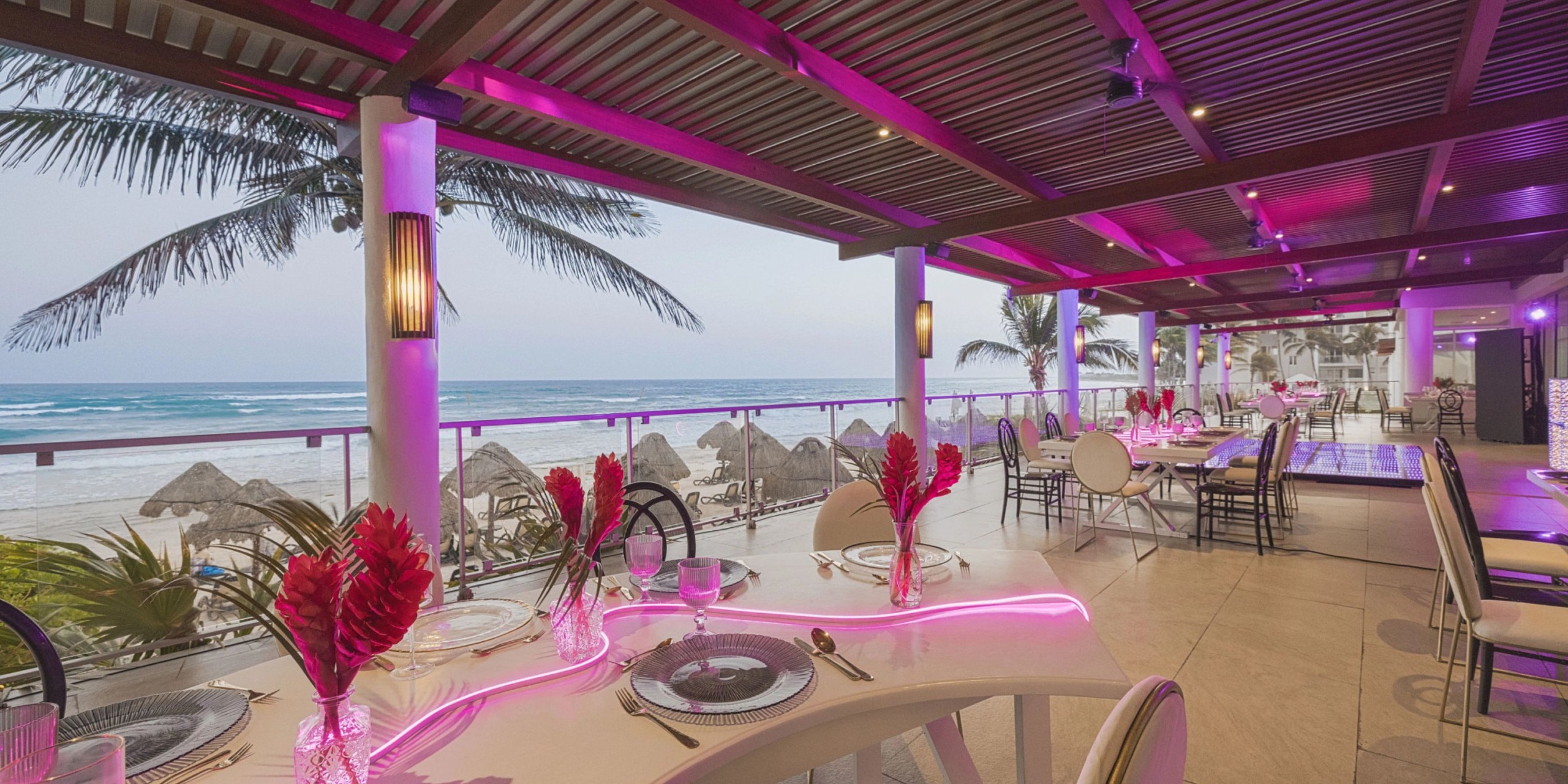 Dreams Jade Riviera Cancun offers a beachfront dining experience with pink lighting, elegantly set tables, red floral centerpieces, and stunning ocean views under a charming wooden canopy.