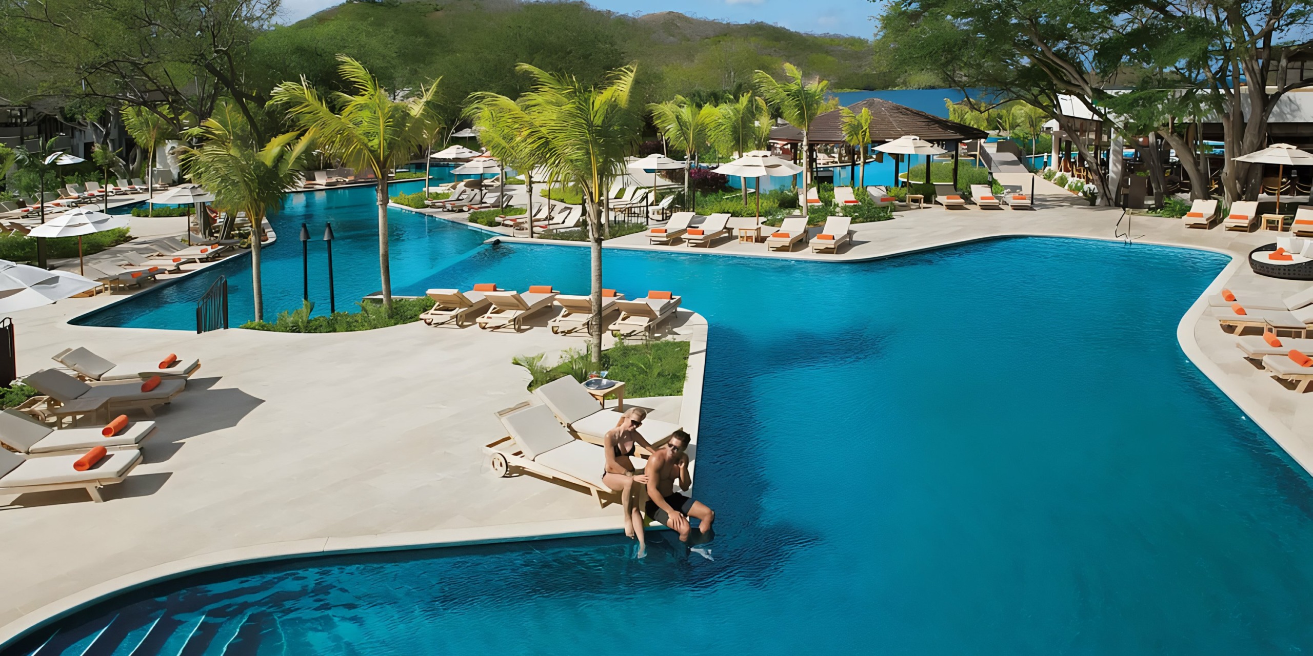 A Dreams Las Mareas resort pool lined with lounge chairs and parasols, with guests relaxing in the sun.