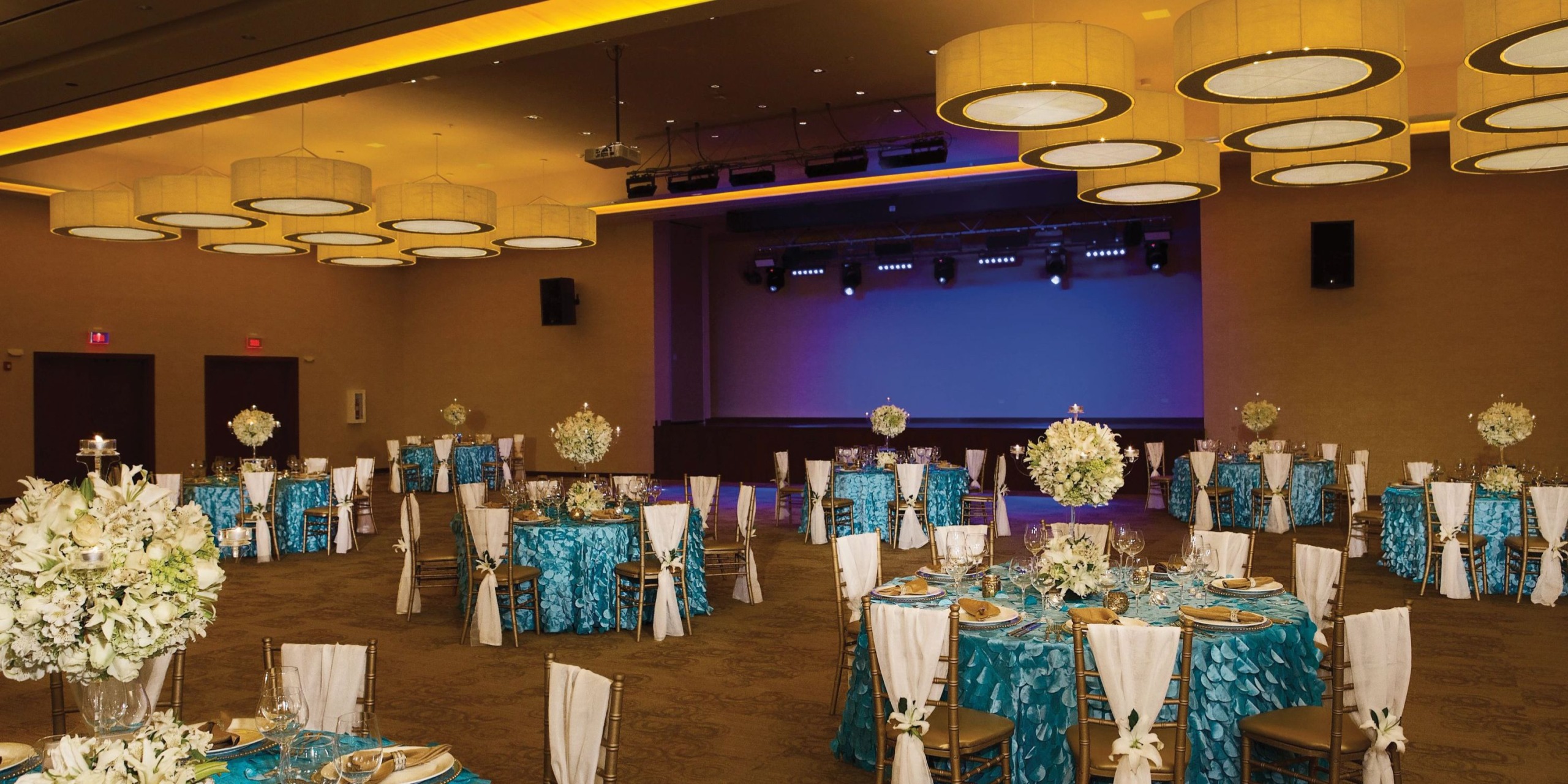 The spacious Dreams Las Mareas banquet hall is adorned with tables featuring teal tablecloths and elegant white floral centerpieces. Overhead, large round ceiling lights illuminate the space, and a stage is prominently situated at the back of the room.