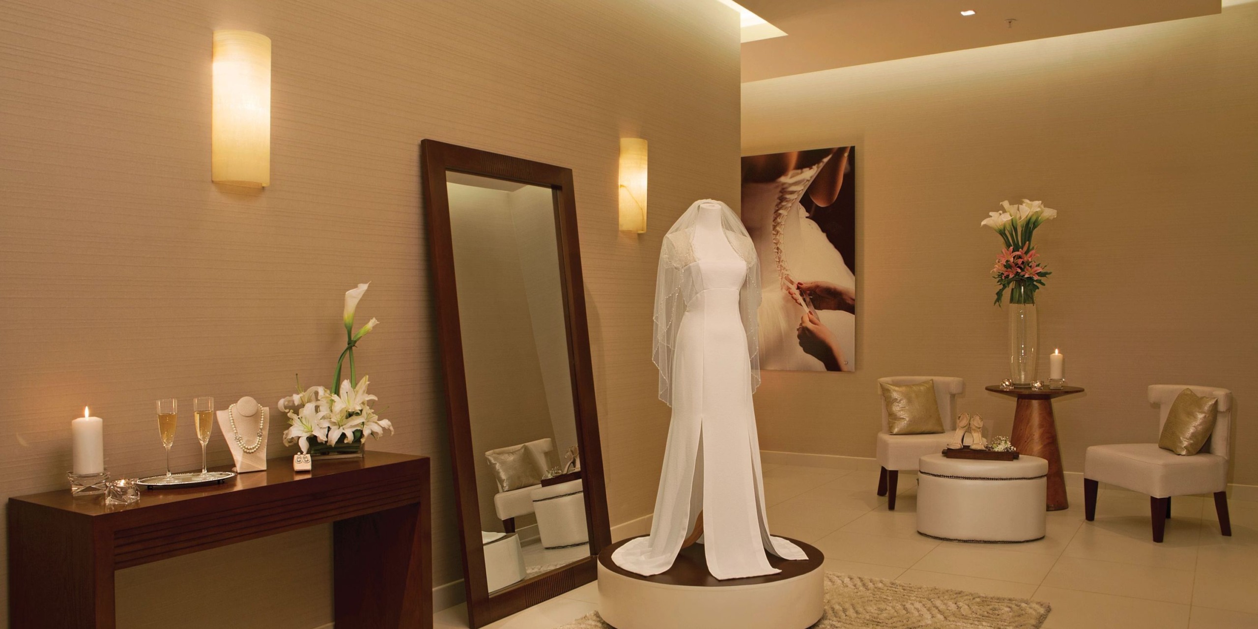 A bridal boutique interior featuring a Dreams Las Mareas floor-length mirror, showcasing a wedding dress on display, with a table adorned with flowers and candles, and seating accompanied by a side table.