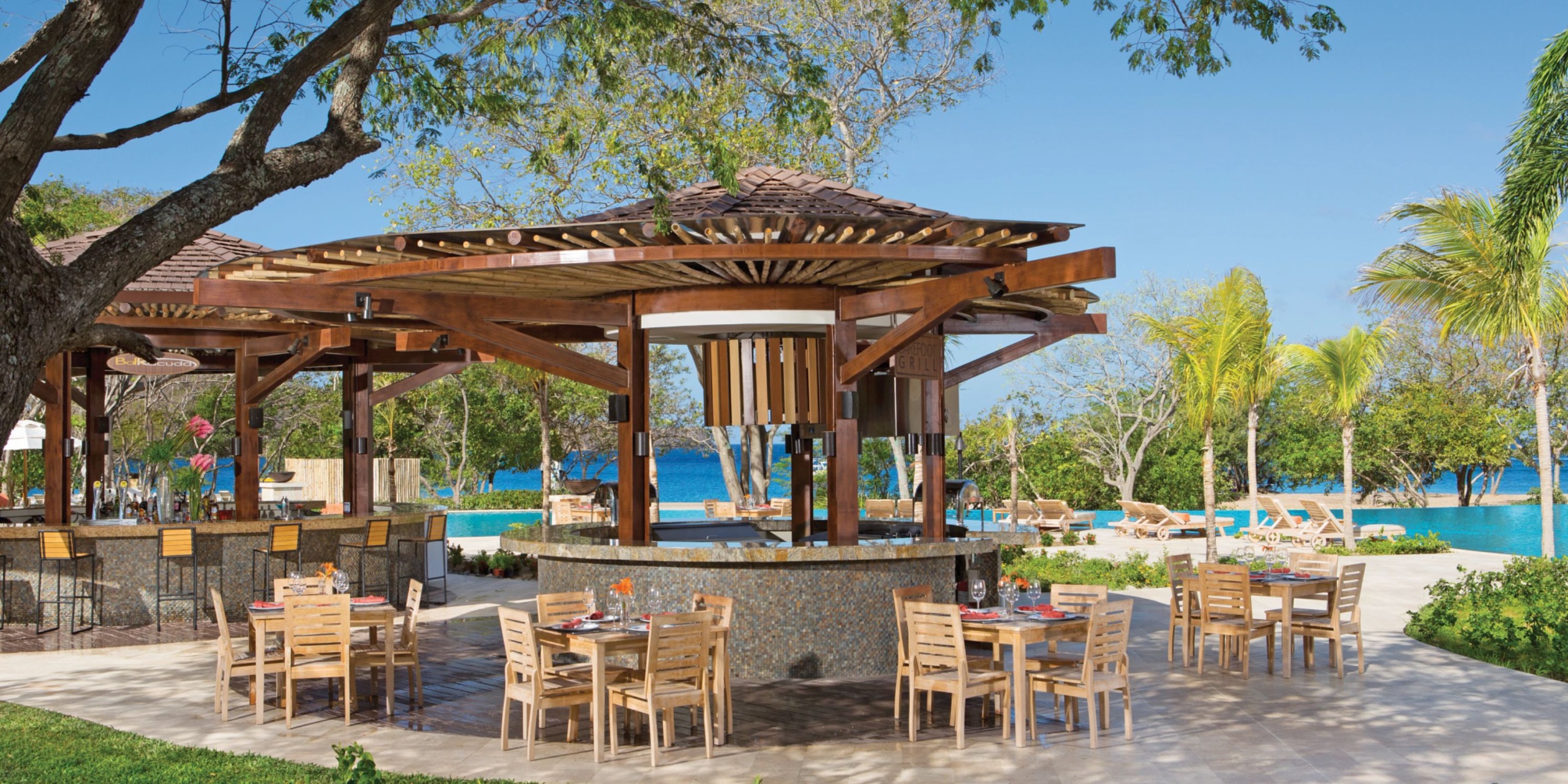 Dining at Dreams Las Mareas involves enjoying wooden tables and chairs under a thatched roof in an outdoor setting. This idyllic location is surrounded by lush trees, a pool, and offers stunning views of the ocean in the background.