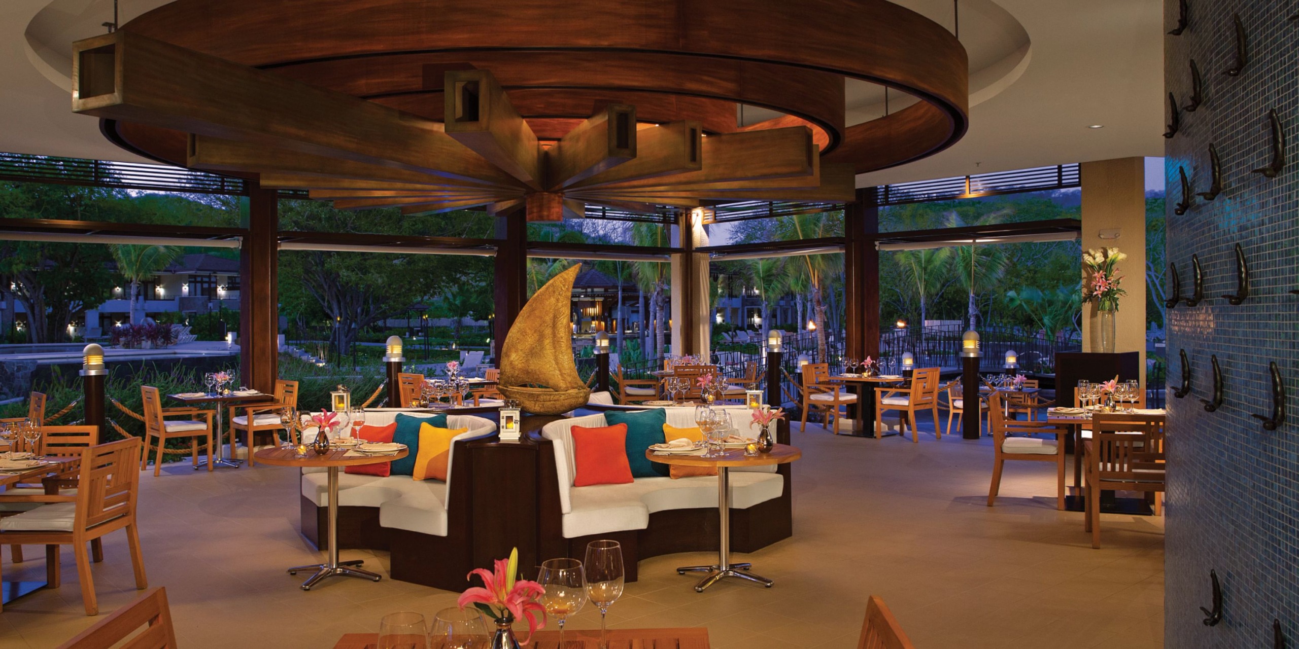 The Dreams Las Mareas features a contemporary restaurant interior adorned with wooden furniture, vibrant cushions, and a striking central sailboat sculpture. Expansive windows provide a scenic view of the lush greenery outside.