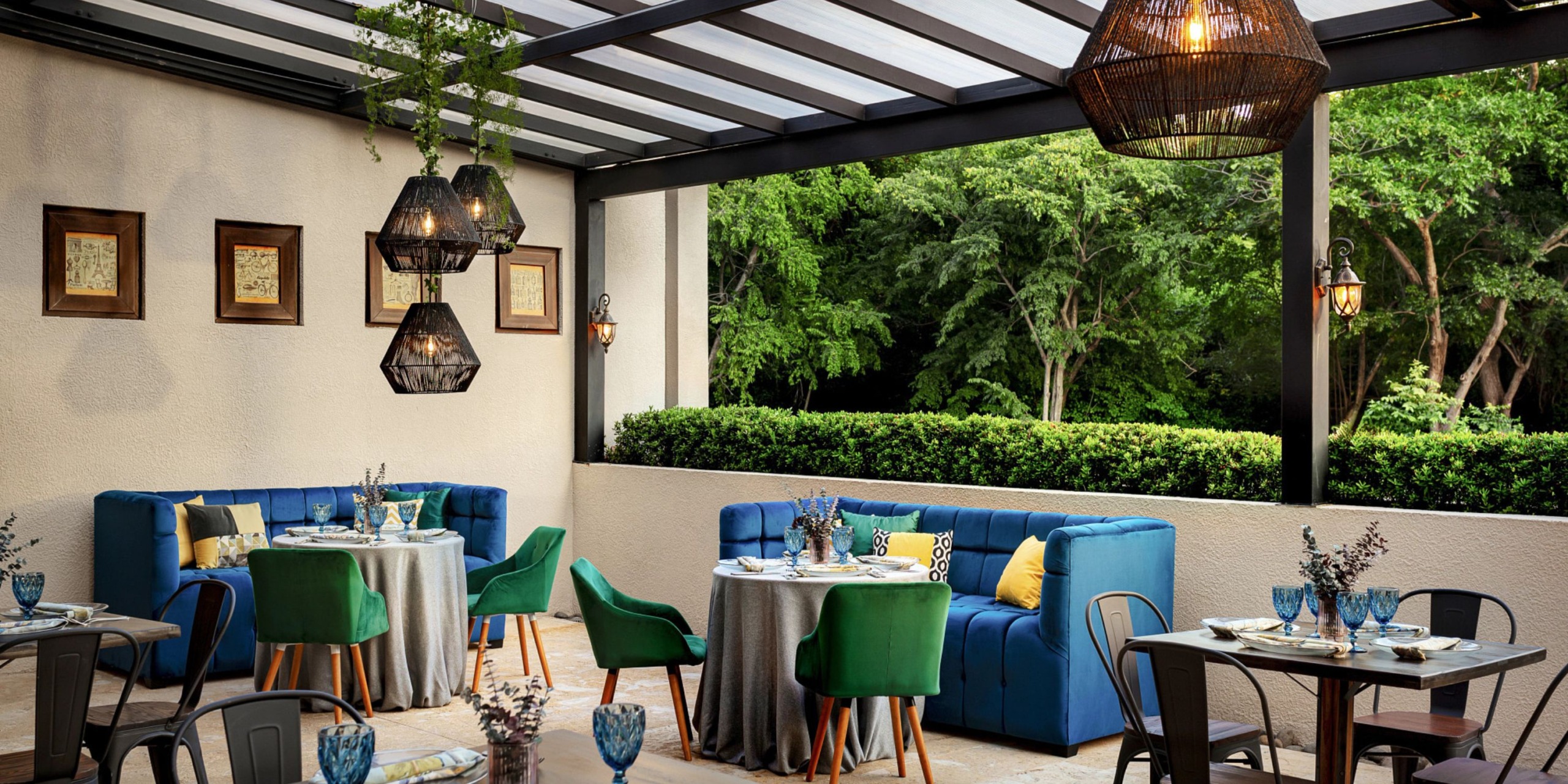 The Dreams Las Mareas features an outdoor patio with blue and green seating, complemented by decorated tables, framed art pieces, and geometric pendant lights, all while offering stunning views of lush greenery.