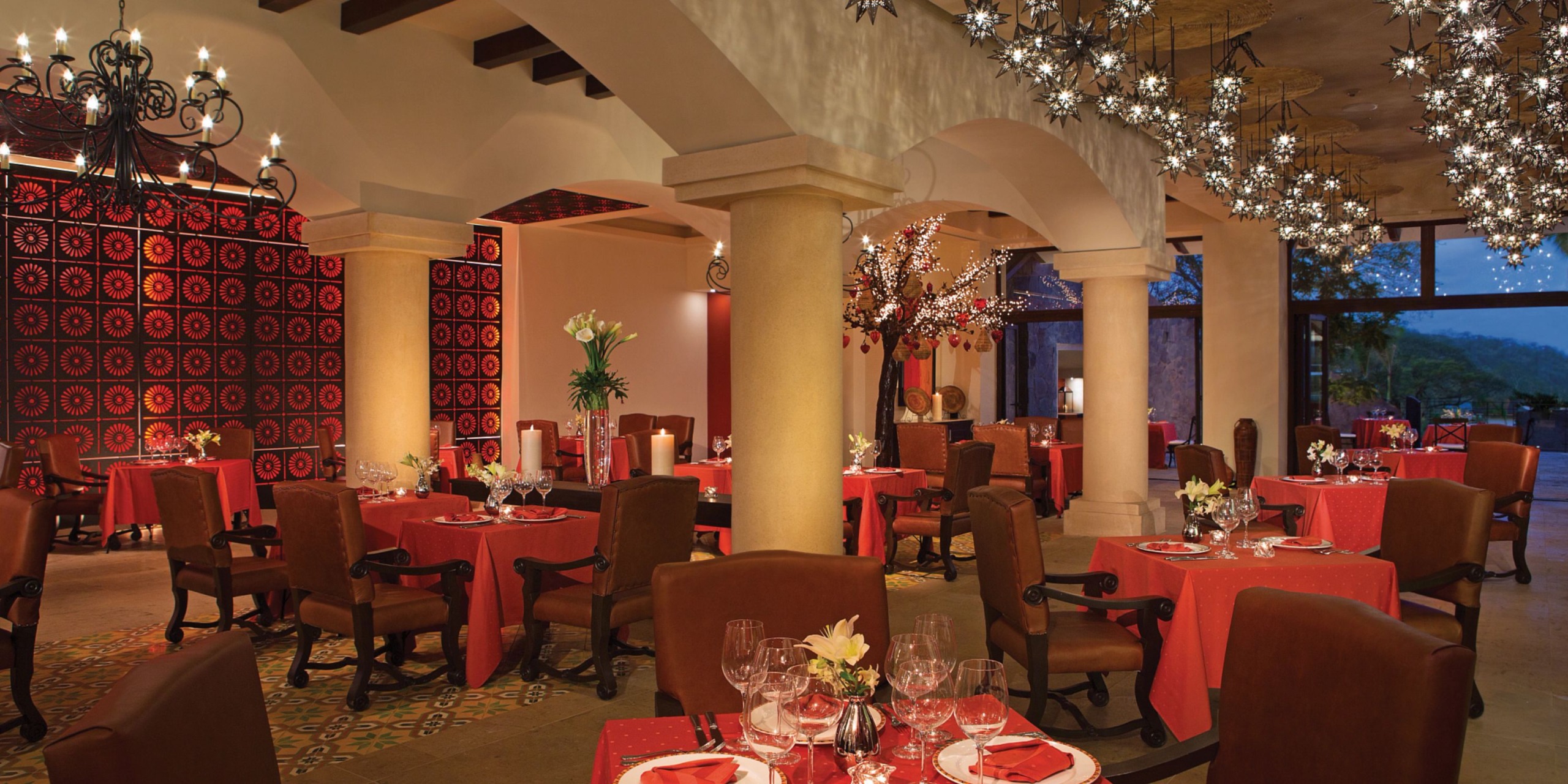 The Dreams Las Mareas offers an elegantly decorated dining space featuring red table settings, ornate chandeliers, and columns, creating a warm and sophisticated atmosphere.