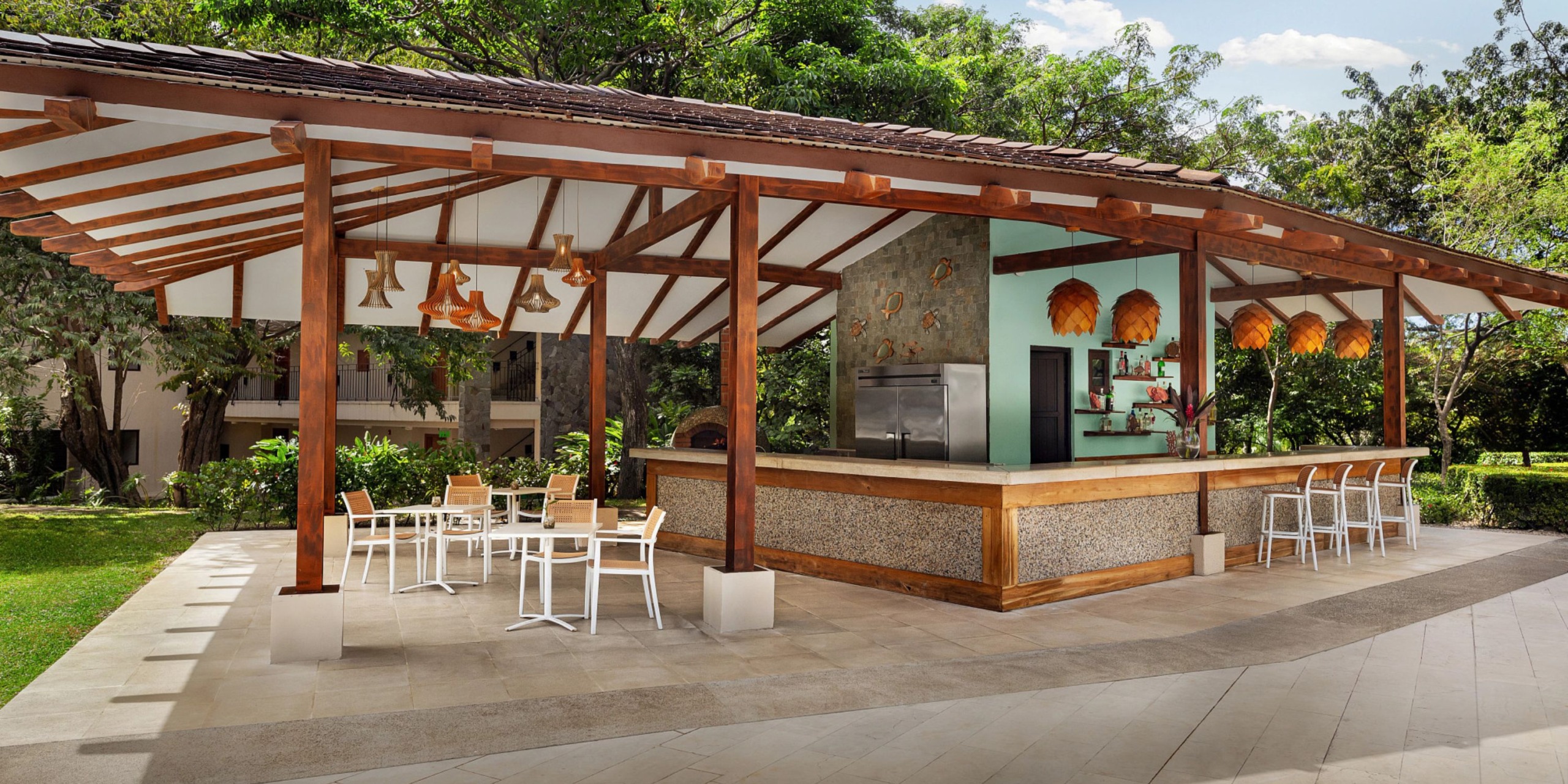 Dreams Las Mareas features an outdoor bar complete with a wooden roof, counter, and stools. The patio is decorated with white tables and chairs set on tiles, surrounded by lush greenery.