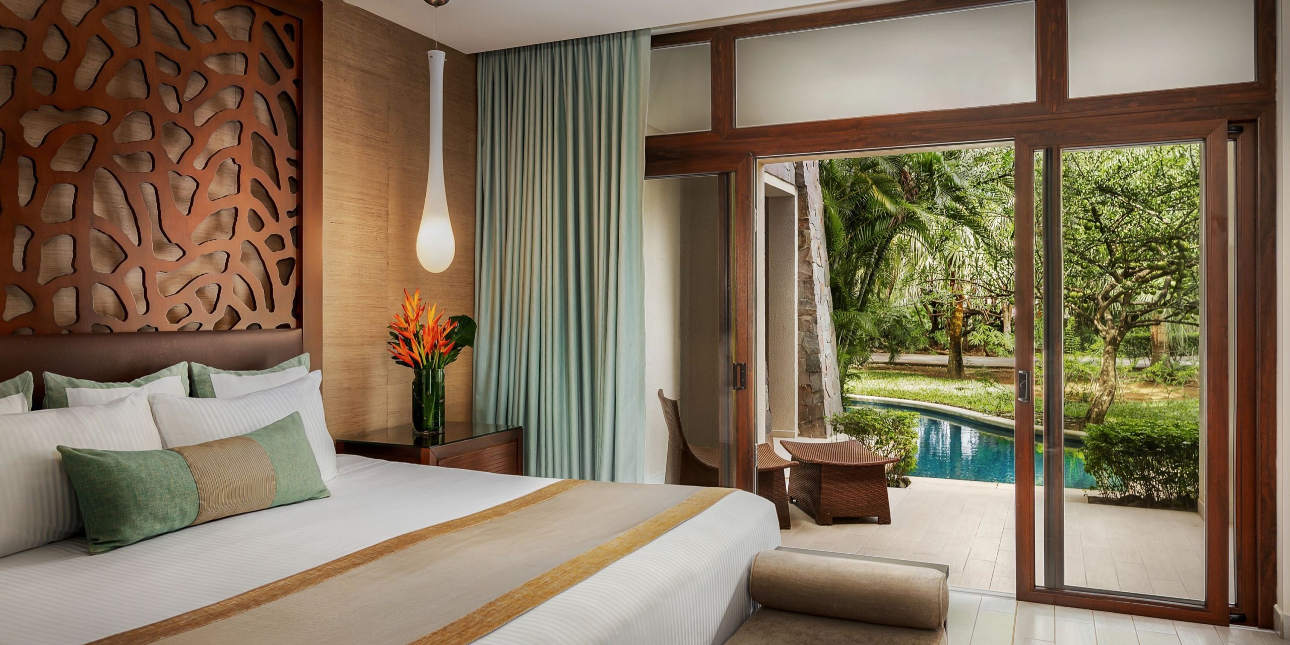 Dreams Las Mareas offers a hotel room featuring a large bed with decorative headboard, complemented by a vase filled with flowers. Glass doors lead to a patio equipped with chairs, overlooking a beautiful pool encircled by trees and lush greenery.