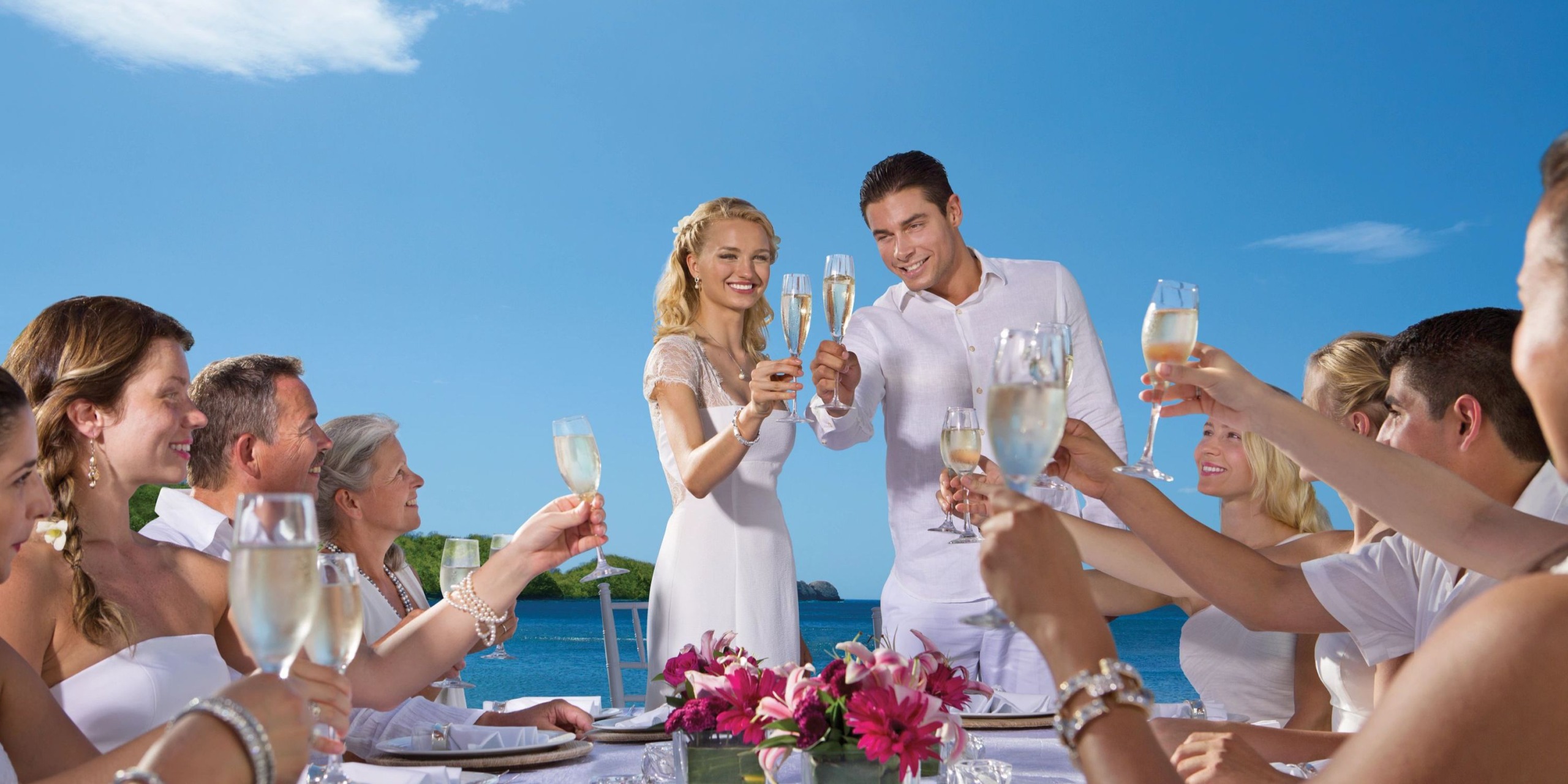 A group of people in white clothing raise their champagne glasses in a toast at Dreams Las Mareas, gathered around a table by the sea under a clear blue sky.