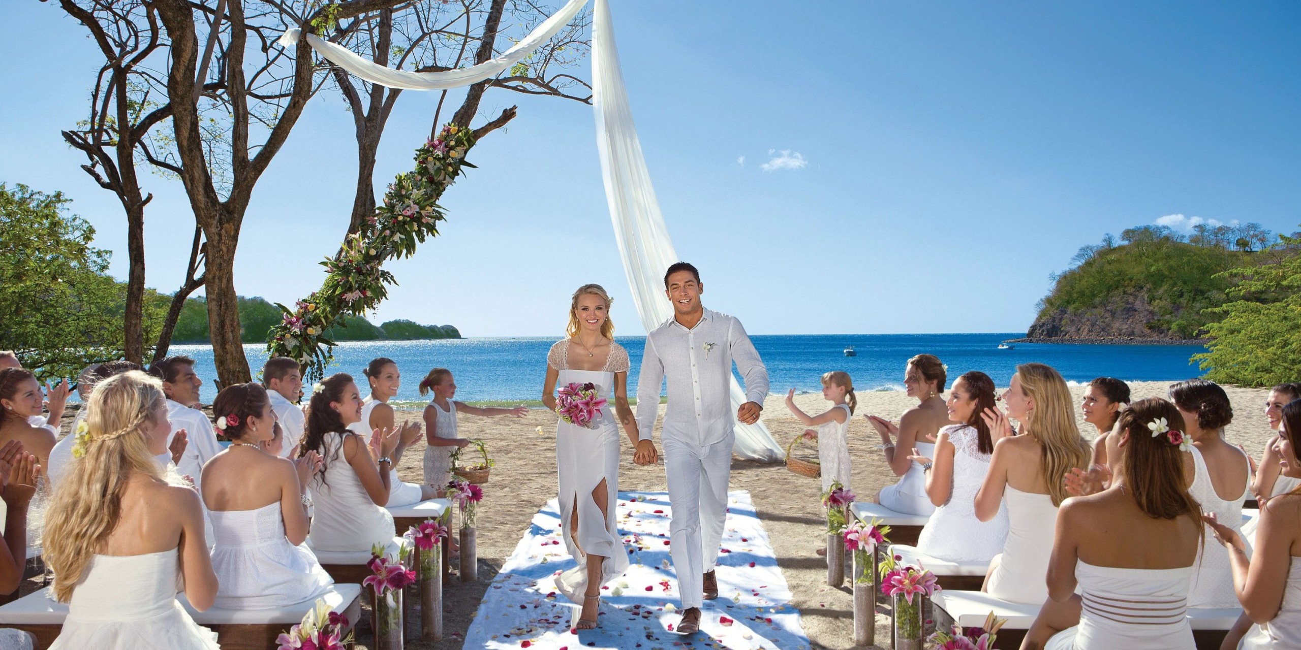 A couple walks hand-in-hand down the aisle at an outdoor beach wedding ceremony at Dreams Las Mareas, surrounded by seated guests dressed in white.