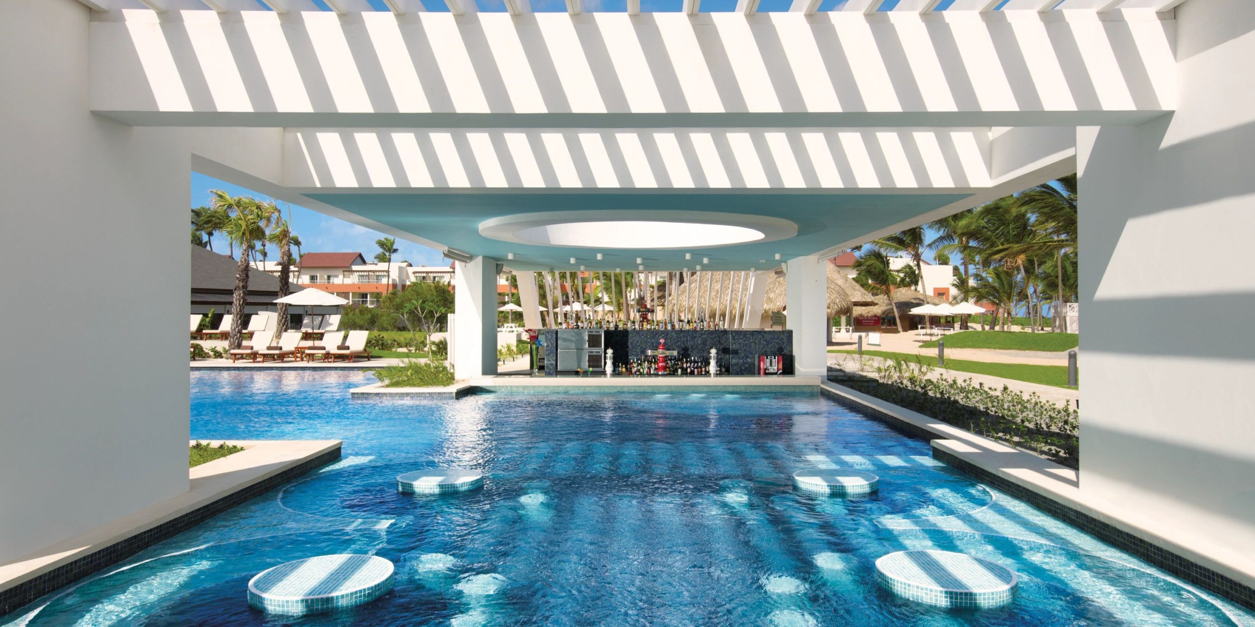 The Dreams Onyx features a poolside bar with submerged seating beneath a contemporary pergola, surrounded by lounge chairs, palm trees, and a resort building in the background.
