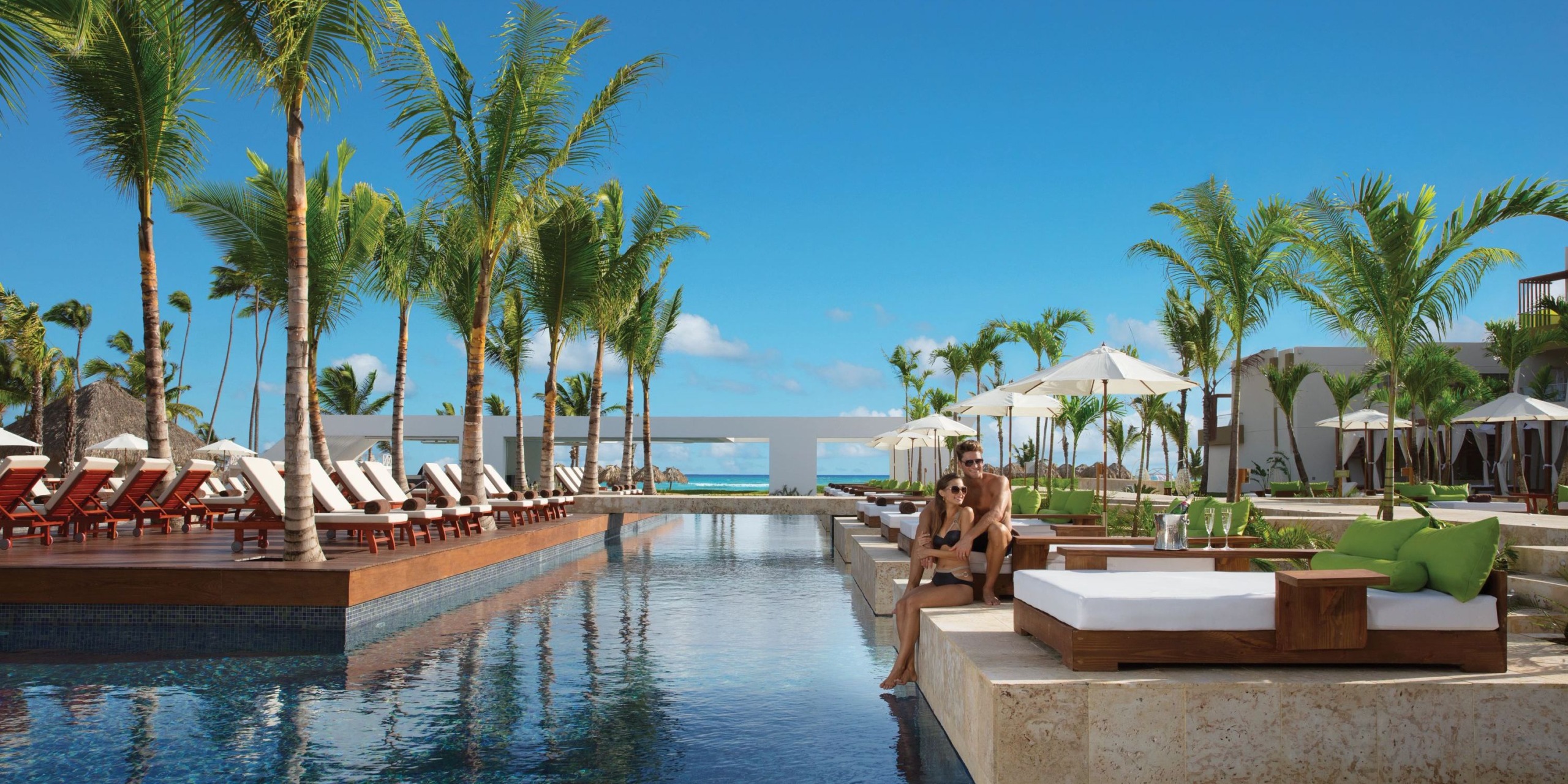 A Dreams Onyx pool area featuring palm trees, lounge chairs, white umbrellas, and two people relaxing on a poolside cushion under a pristine blue sky.