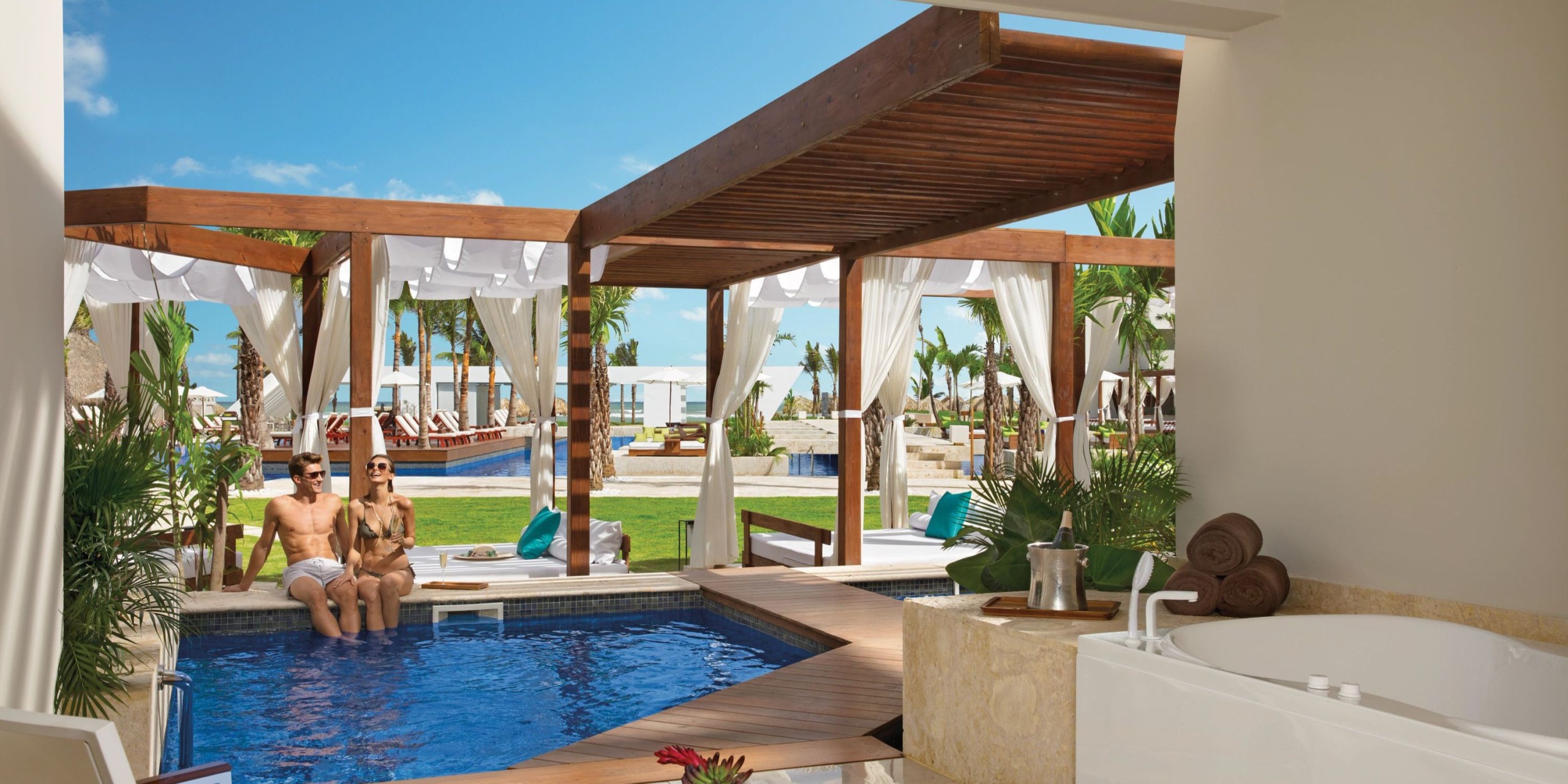 Two people relaxing by a private pool at Dreams Onyx, enjoying the shade of wooden pergolas, with lounge chairs and palm trees in the background.