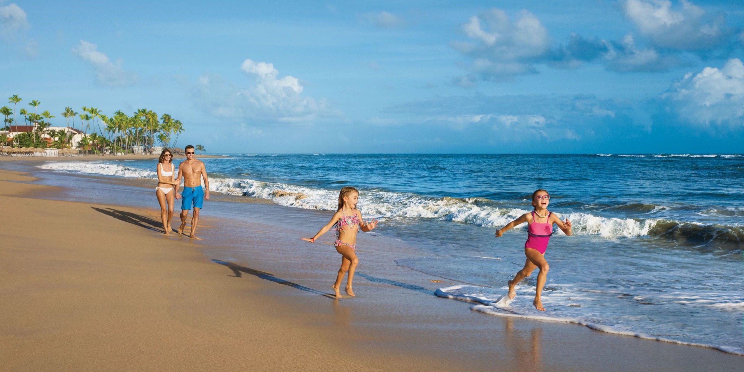 A family strolls along a Dreams Onyx sandy beach with palm trees, as two children enjoy playing near the water under a clear blue sky.