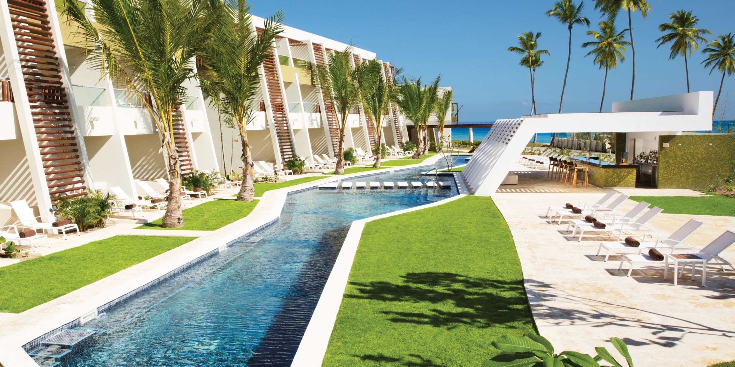 Dreams Onyx features an outdoor resort with a long pool framed by palm trees, lush grass, and comfortable lounge chairs, while a modern bar is conveniently located near the ocean.