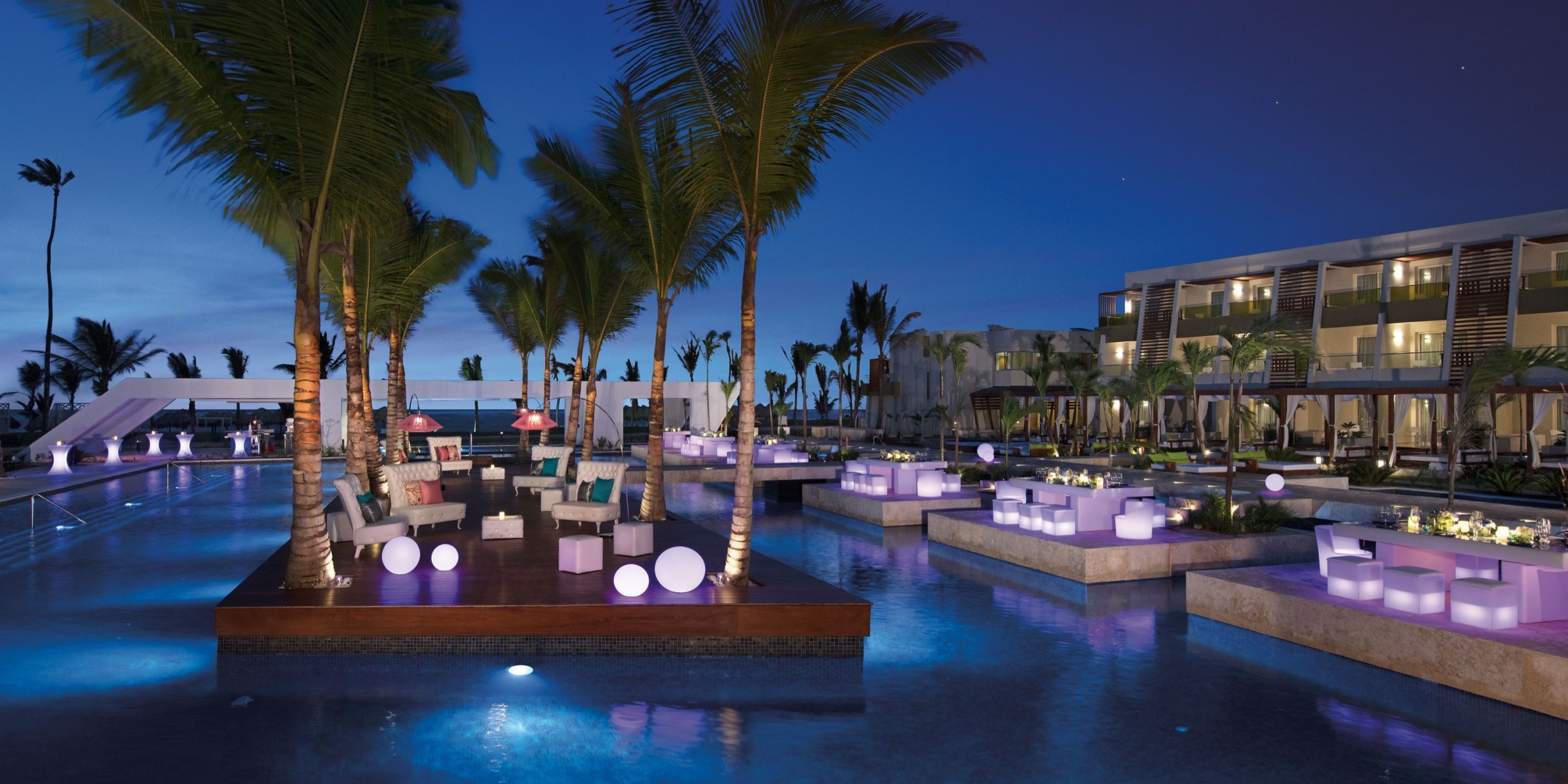 Poolside lounge area at night featuring Dreams Onyx glowing cube and sphere lights, surrounded by palm trees with a nearby modern building.