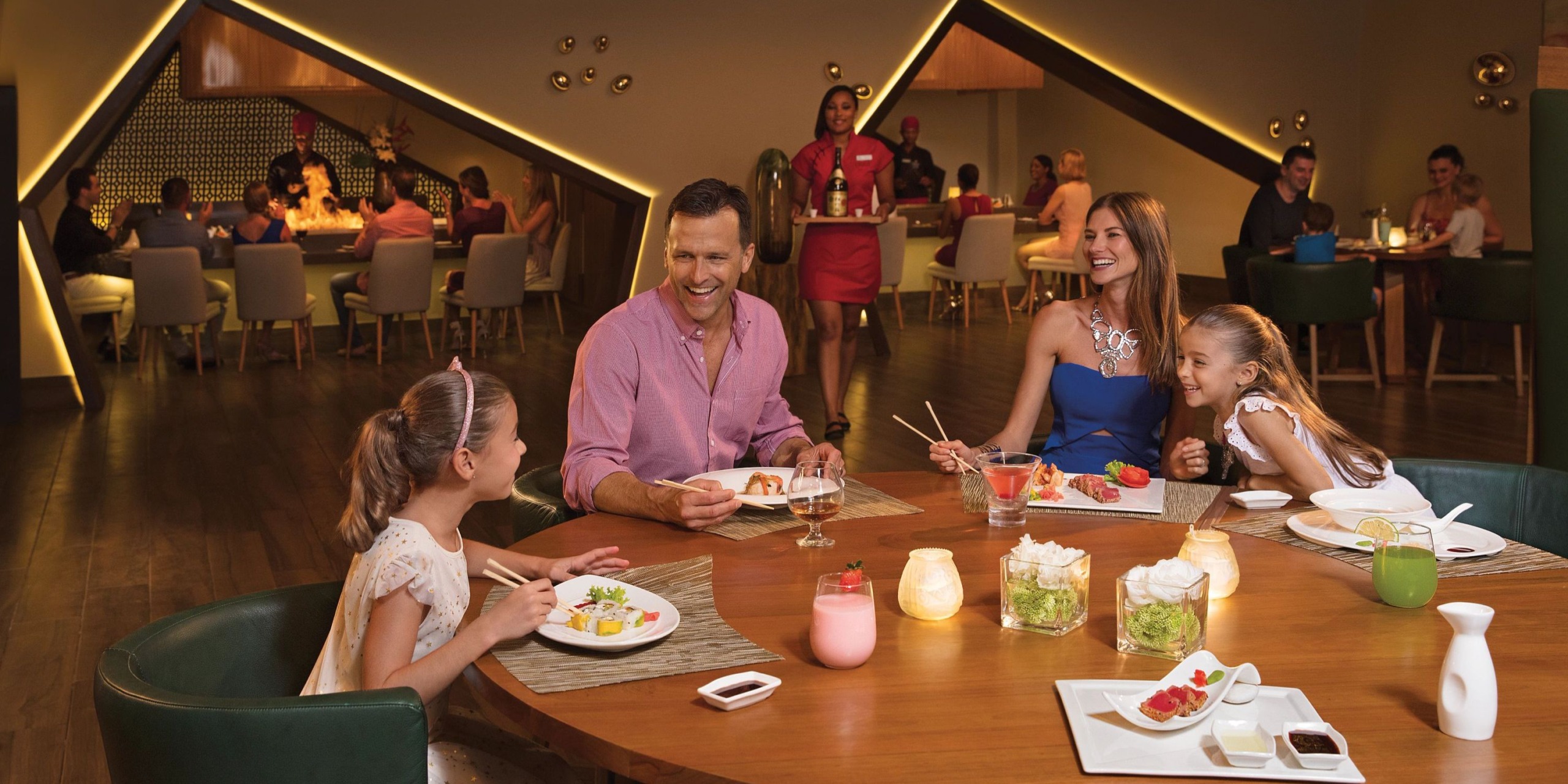 A family of four enjoys a meal together at Dreams Onyx. The table is filled with various dishes and drinks, set in a warmly lit atmosphere with other diners visible in the background.