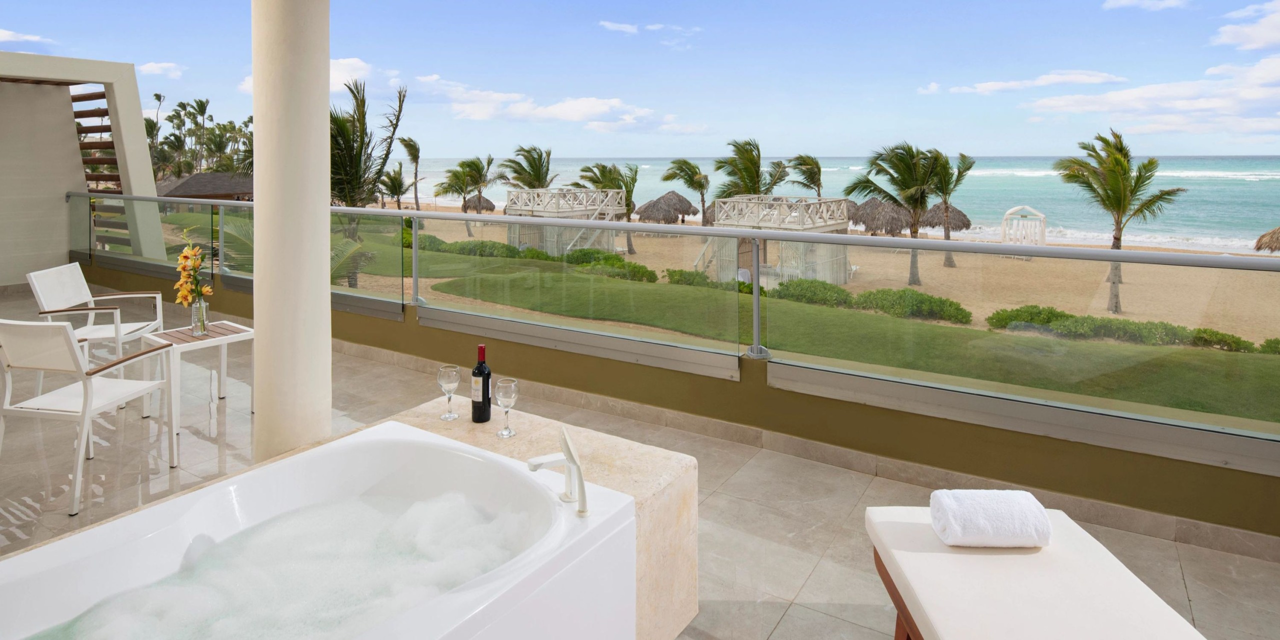 Experience the Dreams Onyx: a beachfront balcony featuring a bubbling hot tub, a cozy table with chairs, a bottle of wine with a glass, all enhanced by the stunning view of palm trees and the ocean.