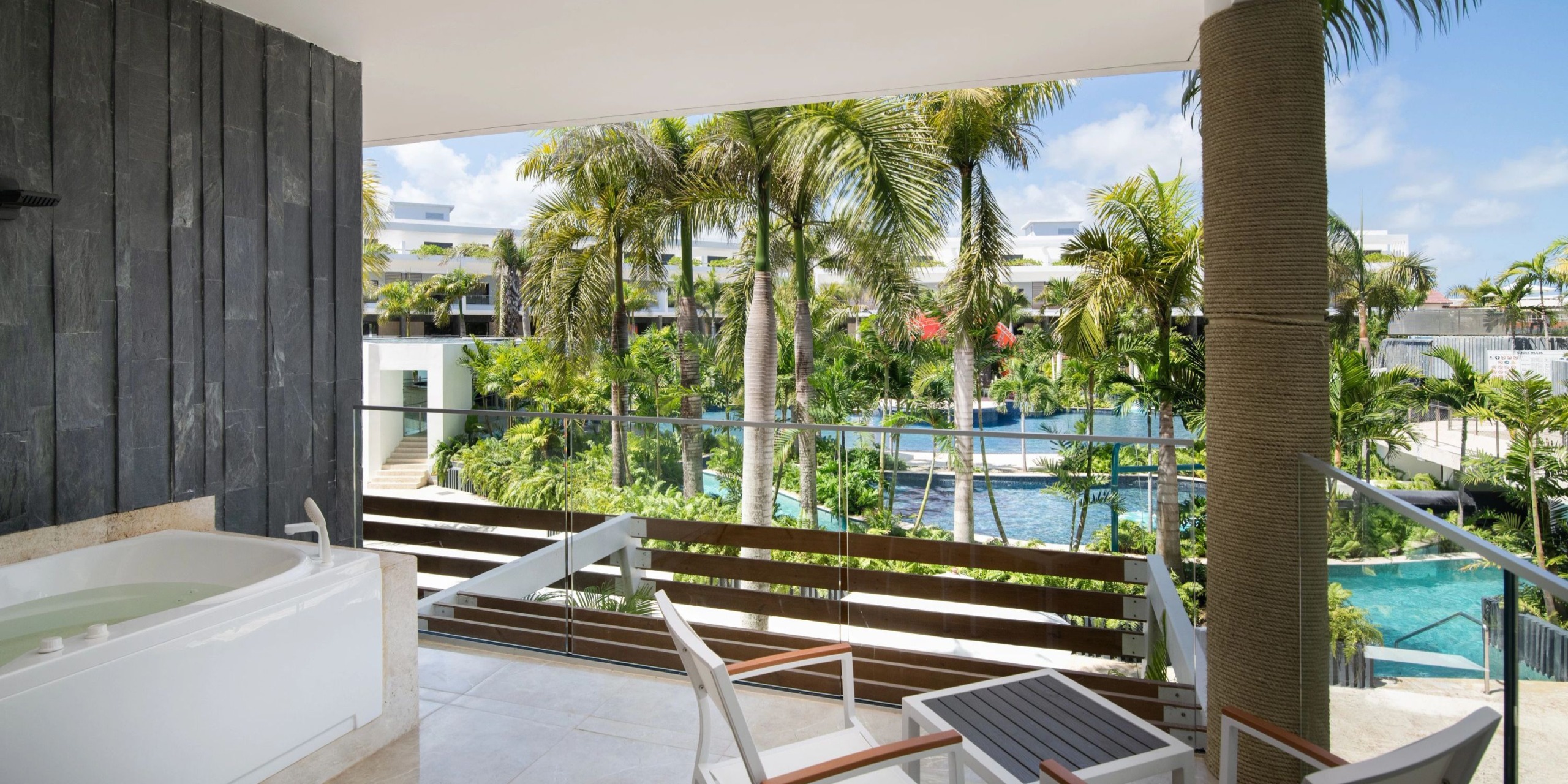 View from the balcony at Dreams Onyx showcasing a tropical resort with palm trees, a swimming pool, and modern architecture. The foreground features a jacuzzi and chairs on the tiled patio.