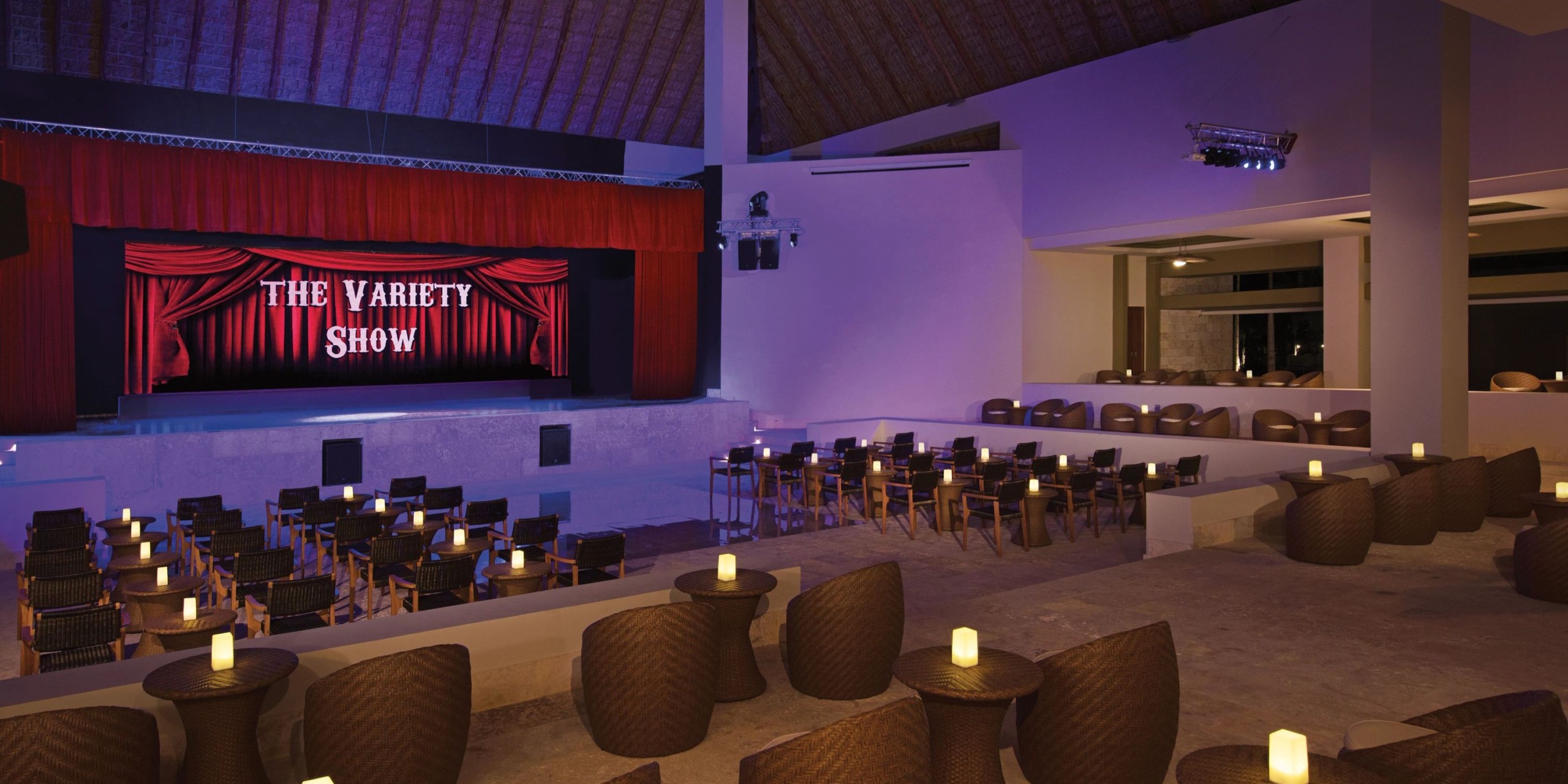 Empty theater with red curtains, a stage, and "Dreams Onyx" on a sign. Rows of chairs and tables with lit candles fill the seating area.