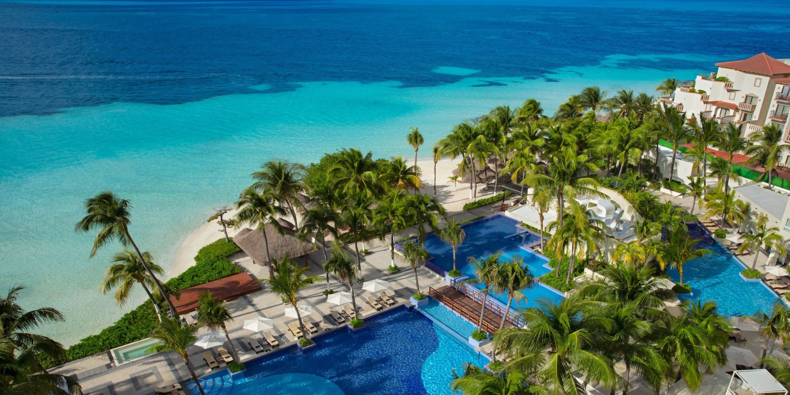 Aerial view of Dreams Sands Cancun, a beachfront resort with multiple pools encircled by palm trees and the turquoise sea shimmering in the background.