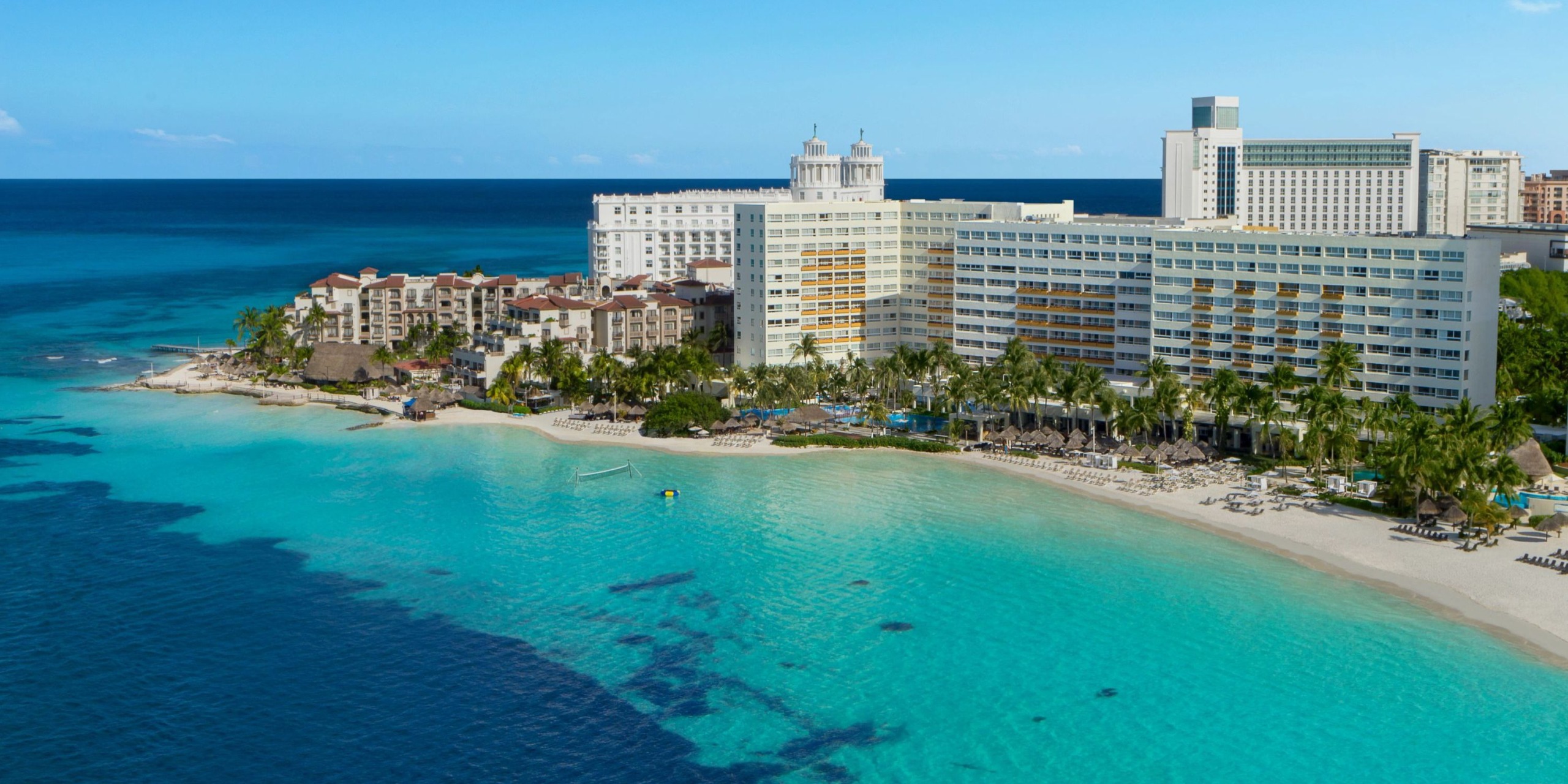 Aerial view of Dreams Sands Cancun, a coastal resort featuring several buildings, palm trees, and a sandy beach by the clear blue ocean under a clear sky.