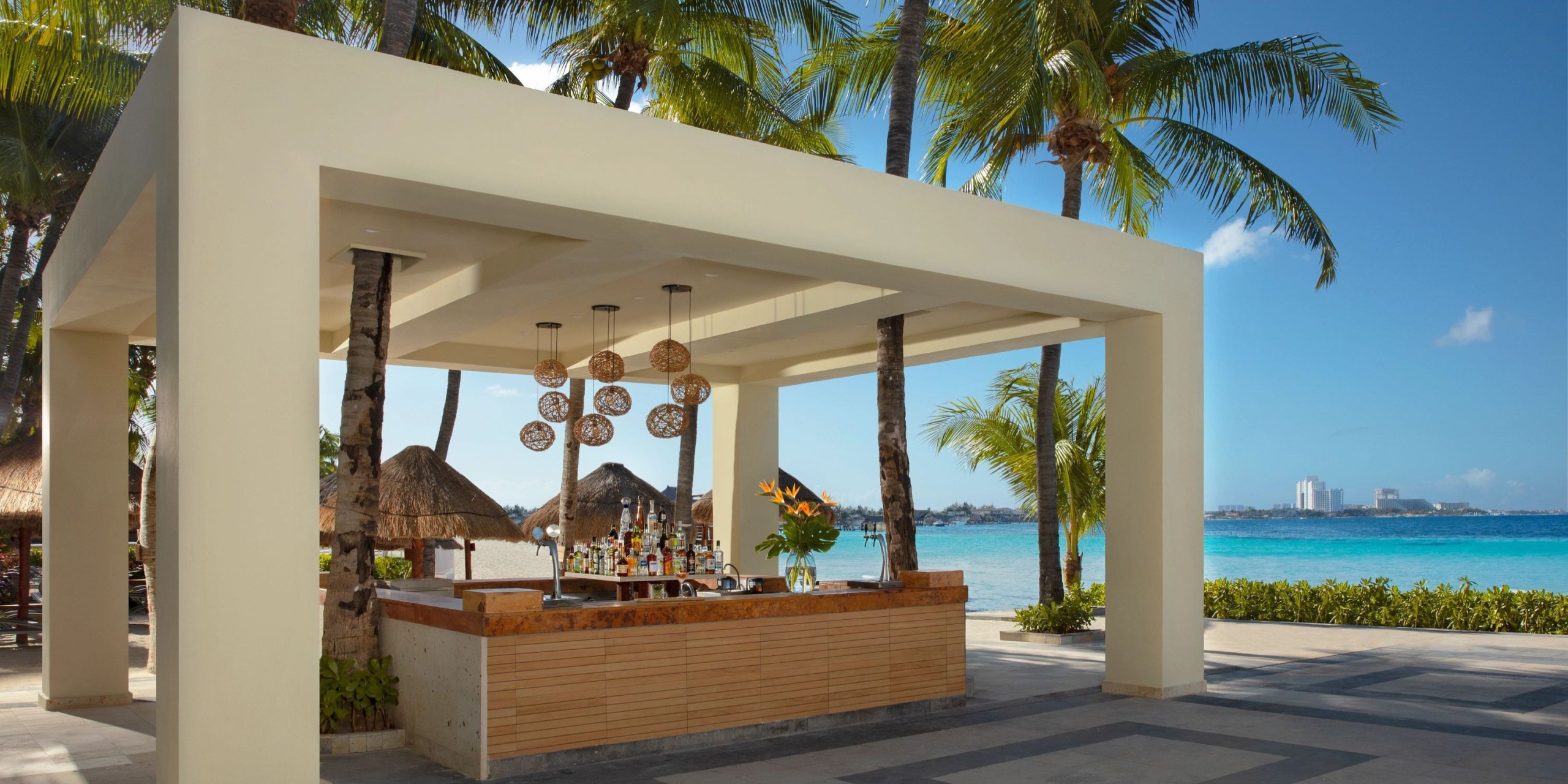 Experience the Dreams Sands Cancun, featuring a beachside bar adorned with hanging lights under a contemporary pergola, nestled amidst palm trees and offering stunning ocean views.