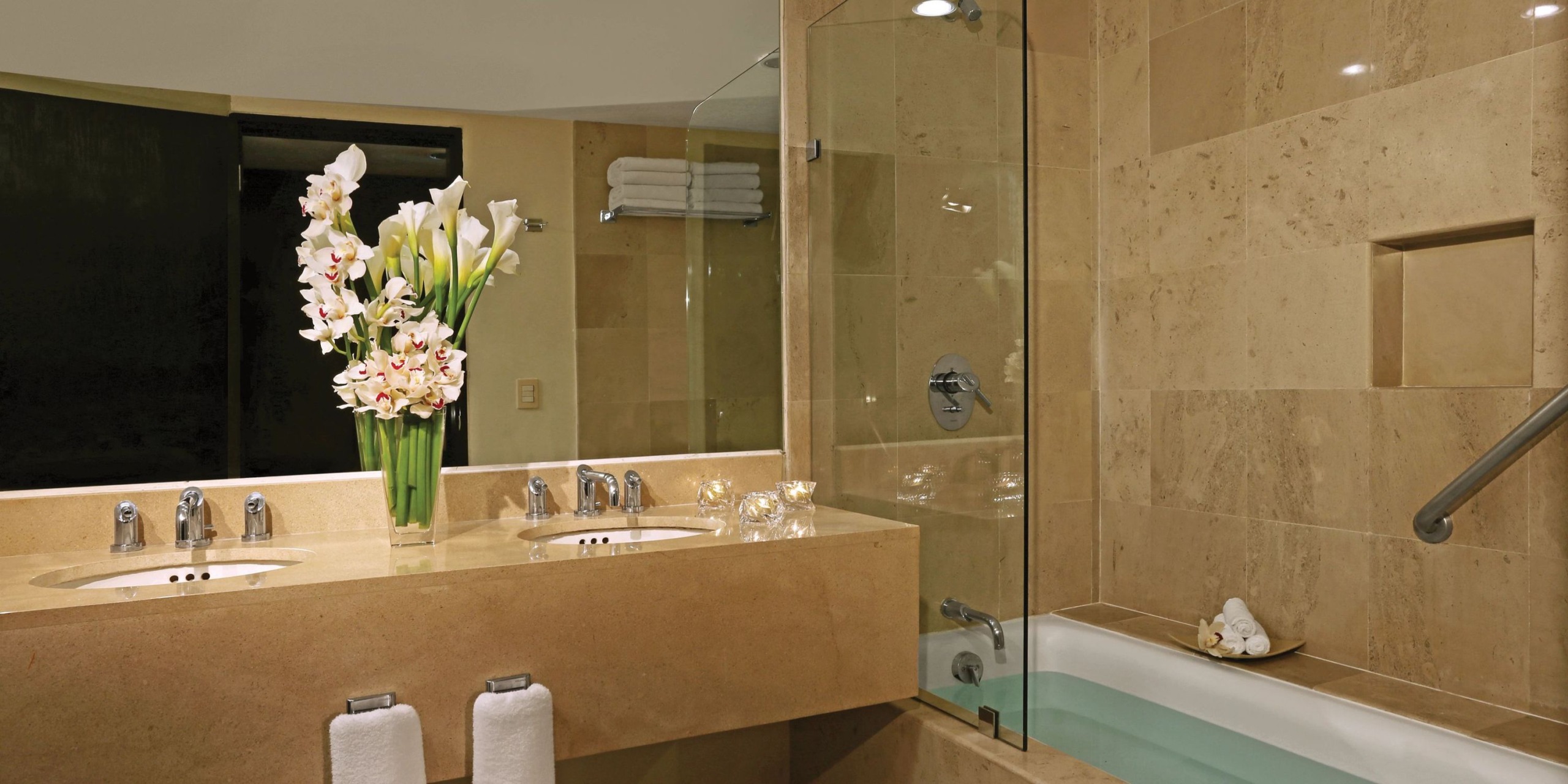 The bathroom at Dreams Sands Cancun features beige tile with a glass-enclosed bathtub, double sinks, and a vase of flowers elegantly placed on the vanity. Towels are conveniently hung below the counter, and neatly folded ones rest on a shelf above.