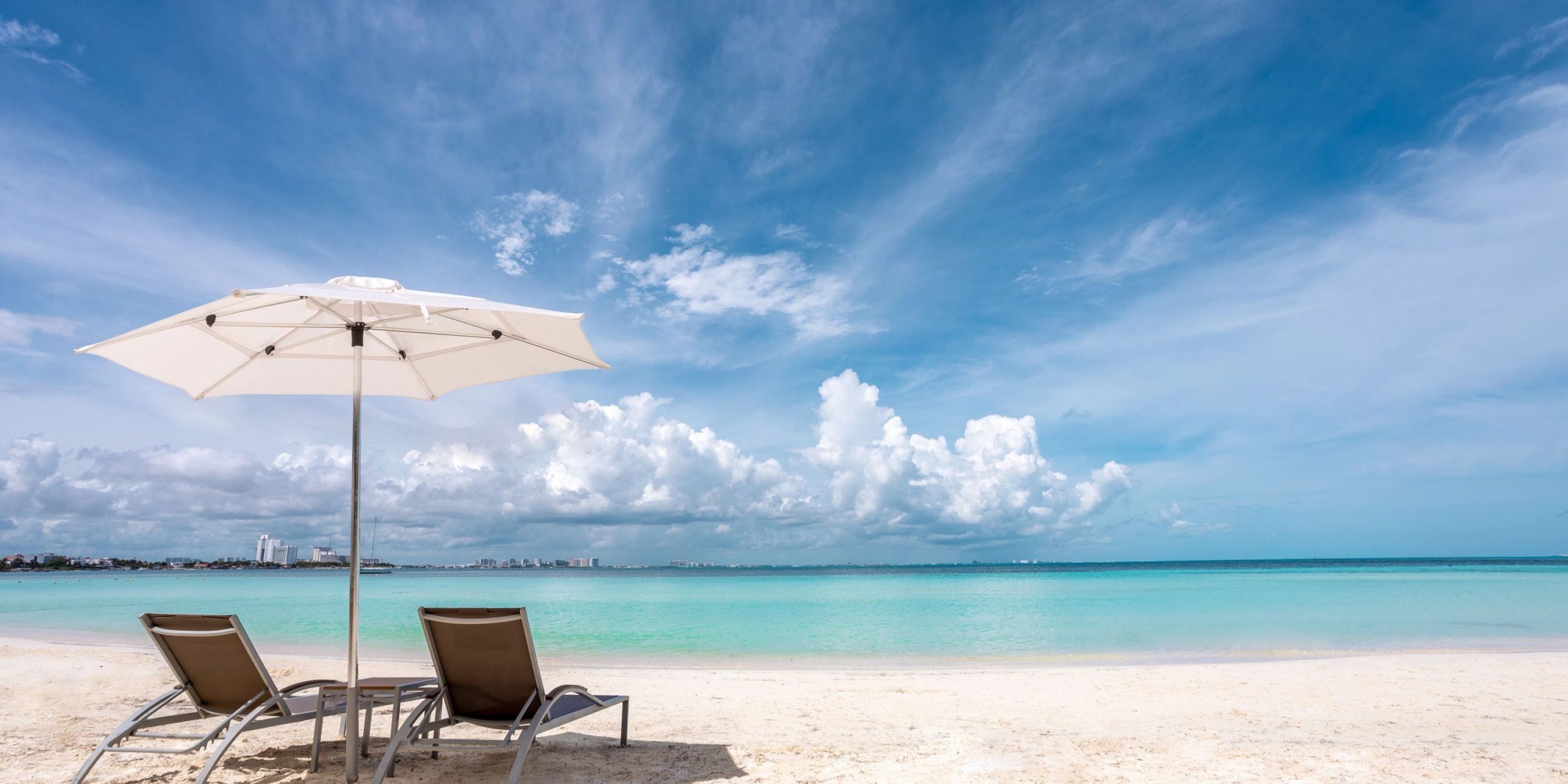 The Dreams Sands Cancun features two lounge chairs shaded by a white umbrella on a sandy beach overlooking the tranquil, turquoise ocean beneath a partly cloudy sky.
