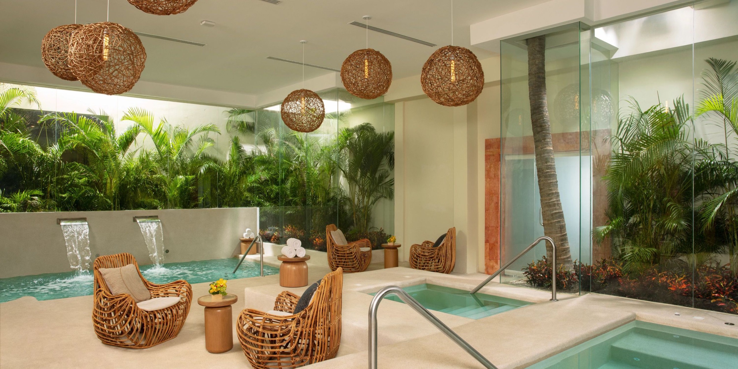 Dreams Sands Cancun features an indoor spa adorned with wicker furniture, a small pool, and tropical plants. Warm lighting is provided by hanging woven lamps, while glass walls offer views of the lush greenery outside.