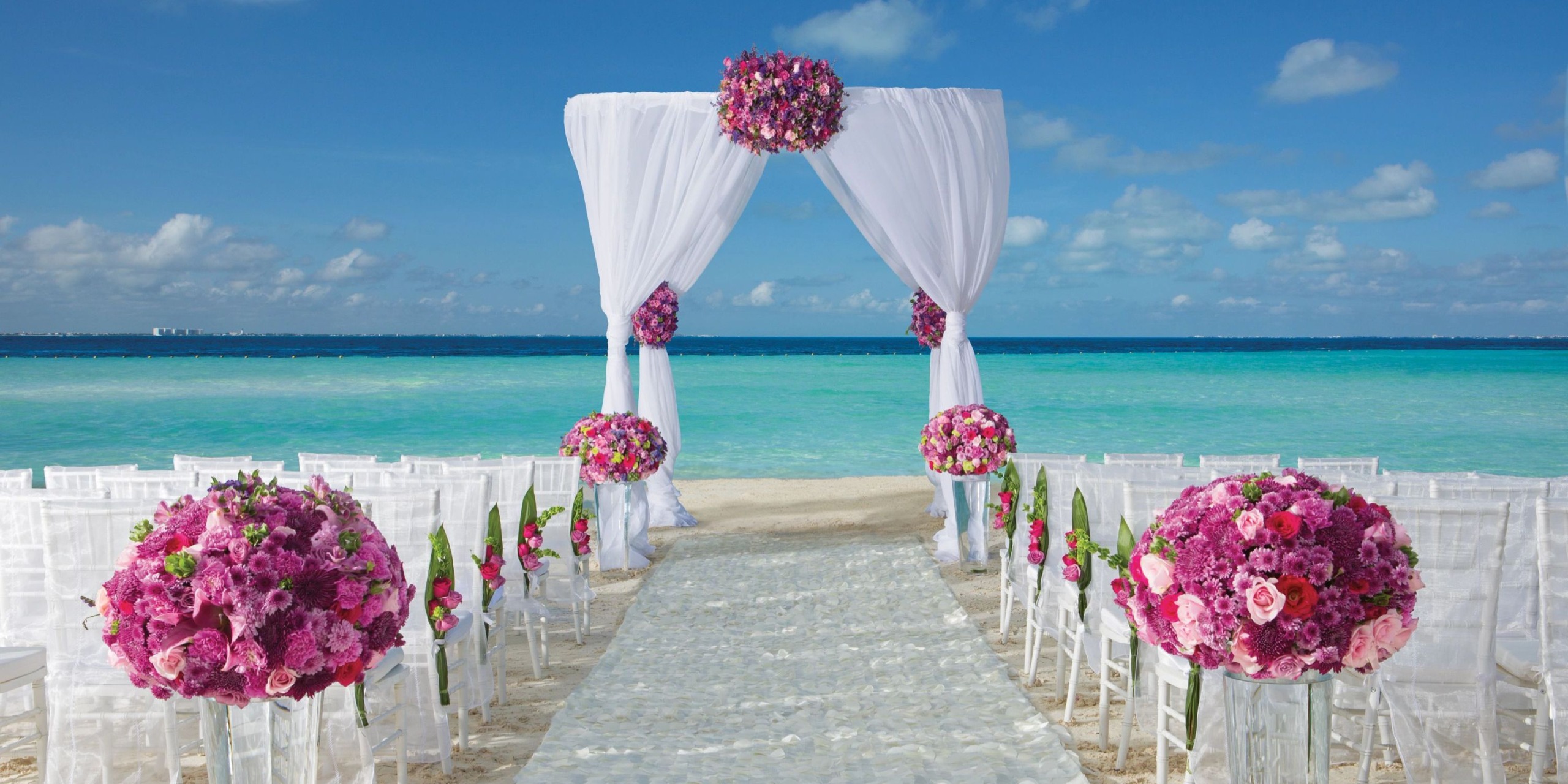Experience a beach wedding setup at Dreams Sands Cancun, featuring a white aisle runner leading to a floral arch, surrounded by white chairs adorned with pink and purple flowers, all overlooking the ocean under a crystal clear blue sky.