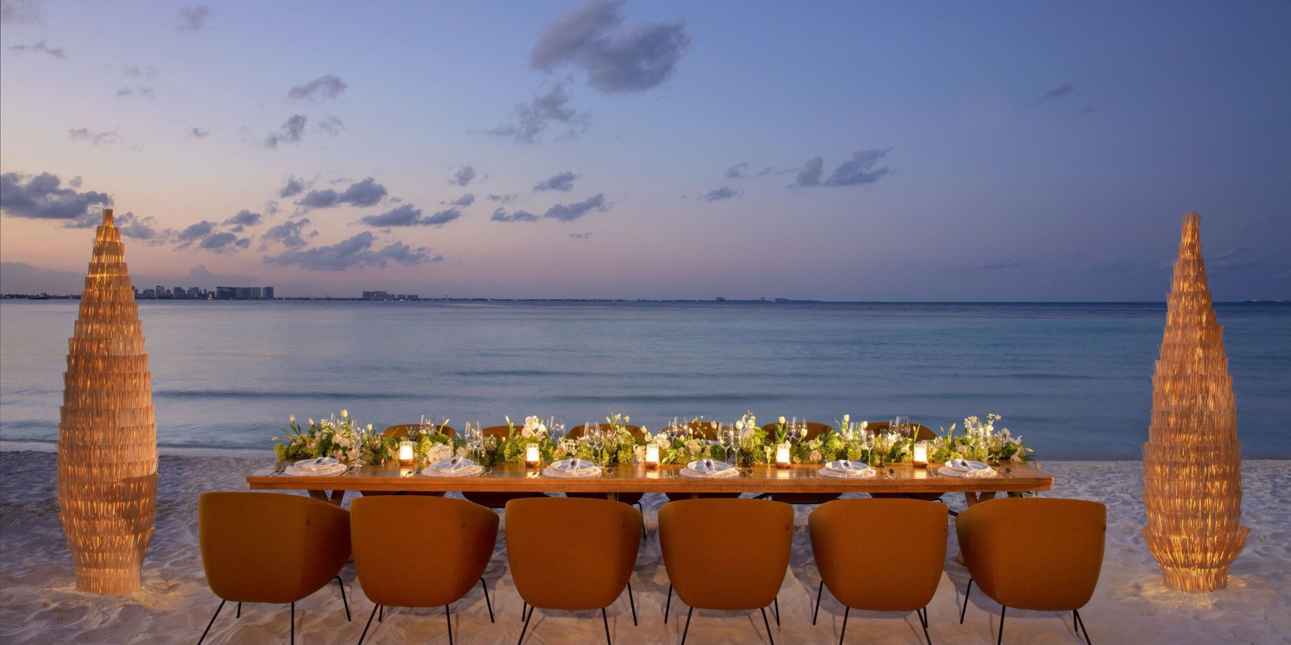 A long Dreams Sands Cancun dining table adorned with floral arrangements is elegantly set on a sandy beach at sunset, with the ocean in the background and clouds gracing the sky.
