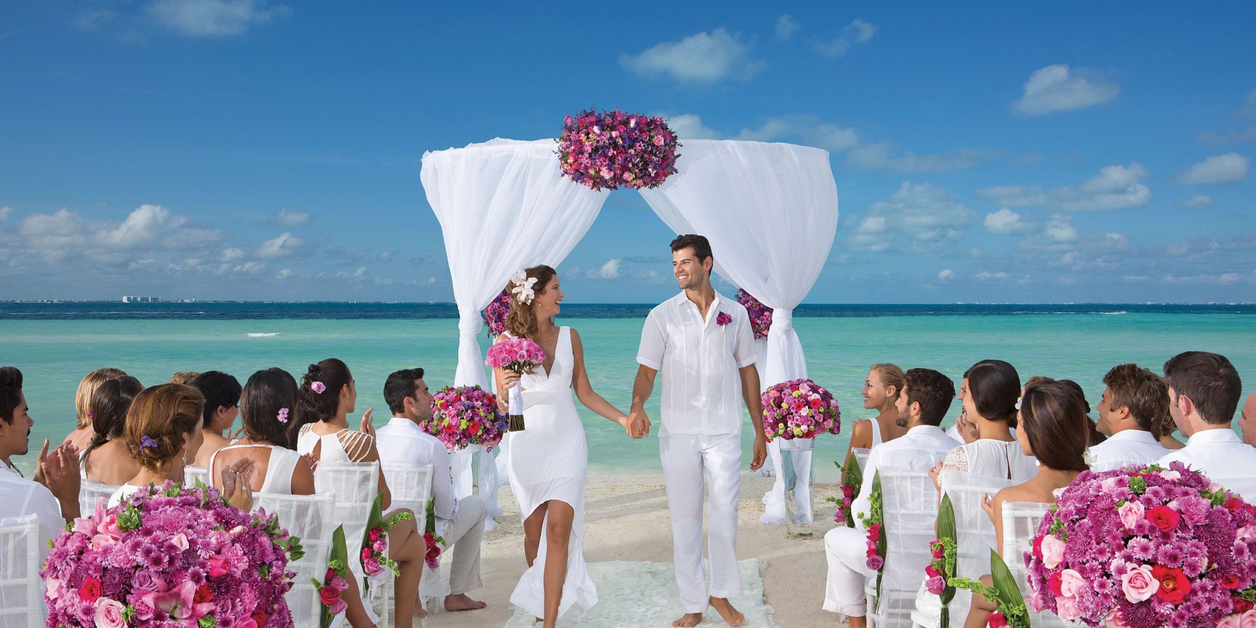 A couple dressed in white is standing under a floral arch on the beach at Dreams Sands Cancun, holding hands and smiling. Guests dressed in white are seated, facing the couple.
