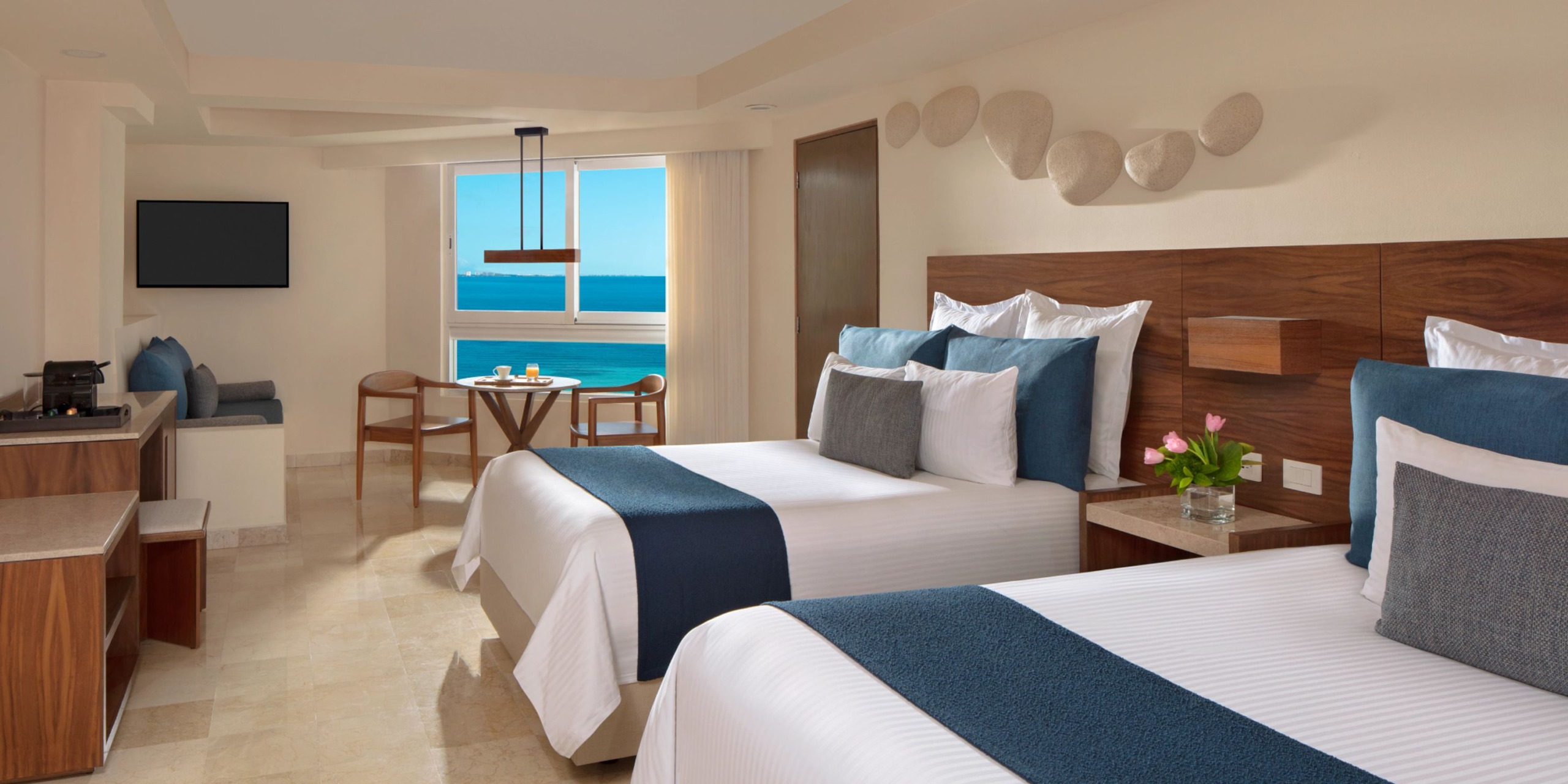 Experience the Dreams Sands Cancun, featuring a hotel room with two double beds, a cozy dining area, a wall-mounted TV, and an ocean-view window. The room is elegantly designed with neutral decor complemented by blue accents.