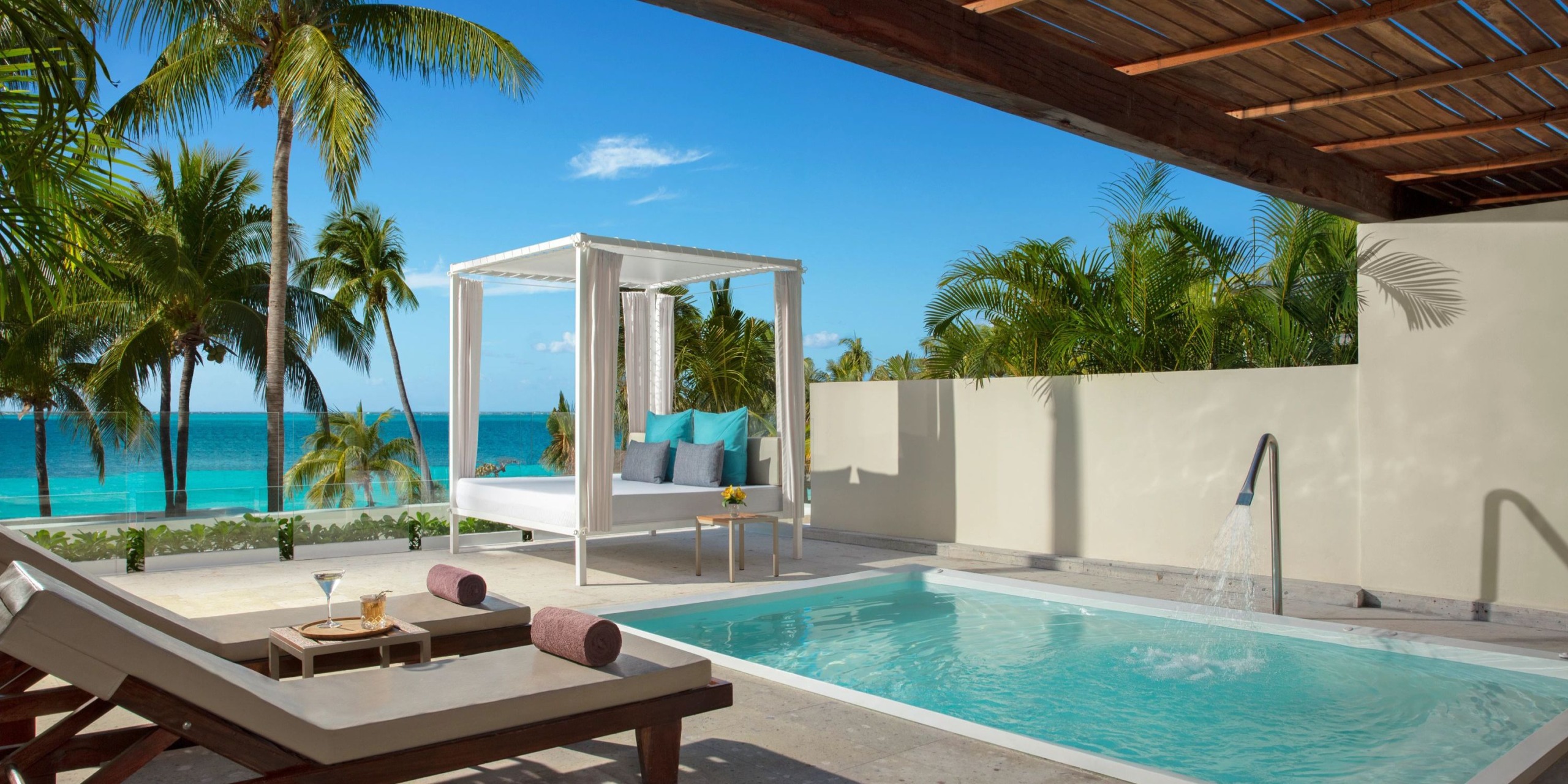 Experience the luxurious poolside area at Dreams Sands Cancun, complete with lounge chairs, a canopy bed, and a stunning ocean view. Enjoy the tropical palm trees and clear blue sky that create an idyllic atmosphere.