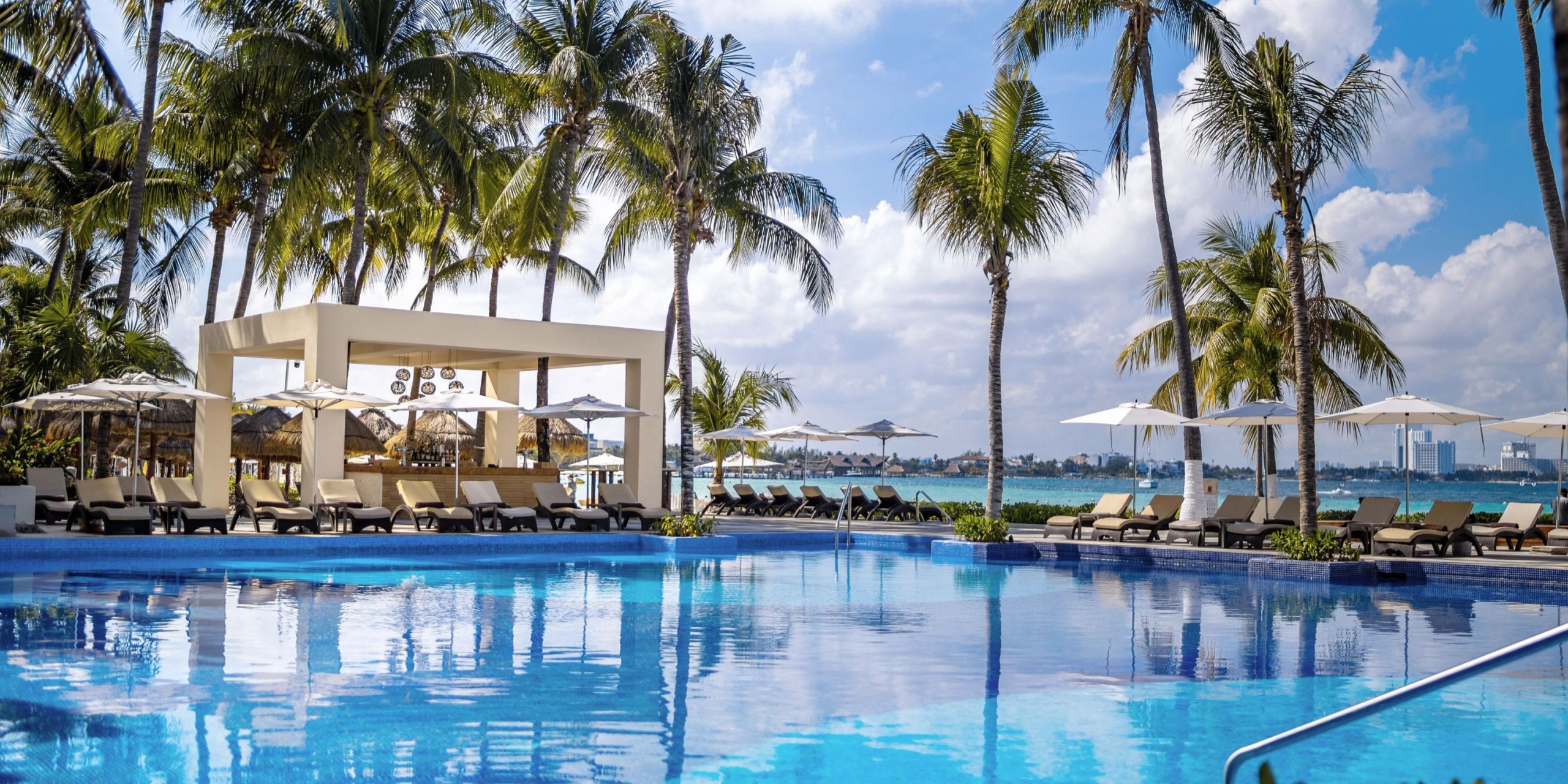 Dreams Sands Cancun offers a picturesque poolside scene with palm trees, sun loungers, and umbrellas beside a clear blue swimming pool. On the left, there's a bar structure overlooking a body of water beneath a partly cloudy sky.