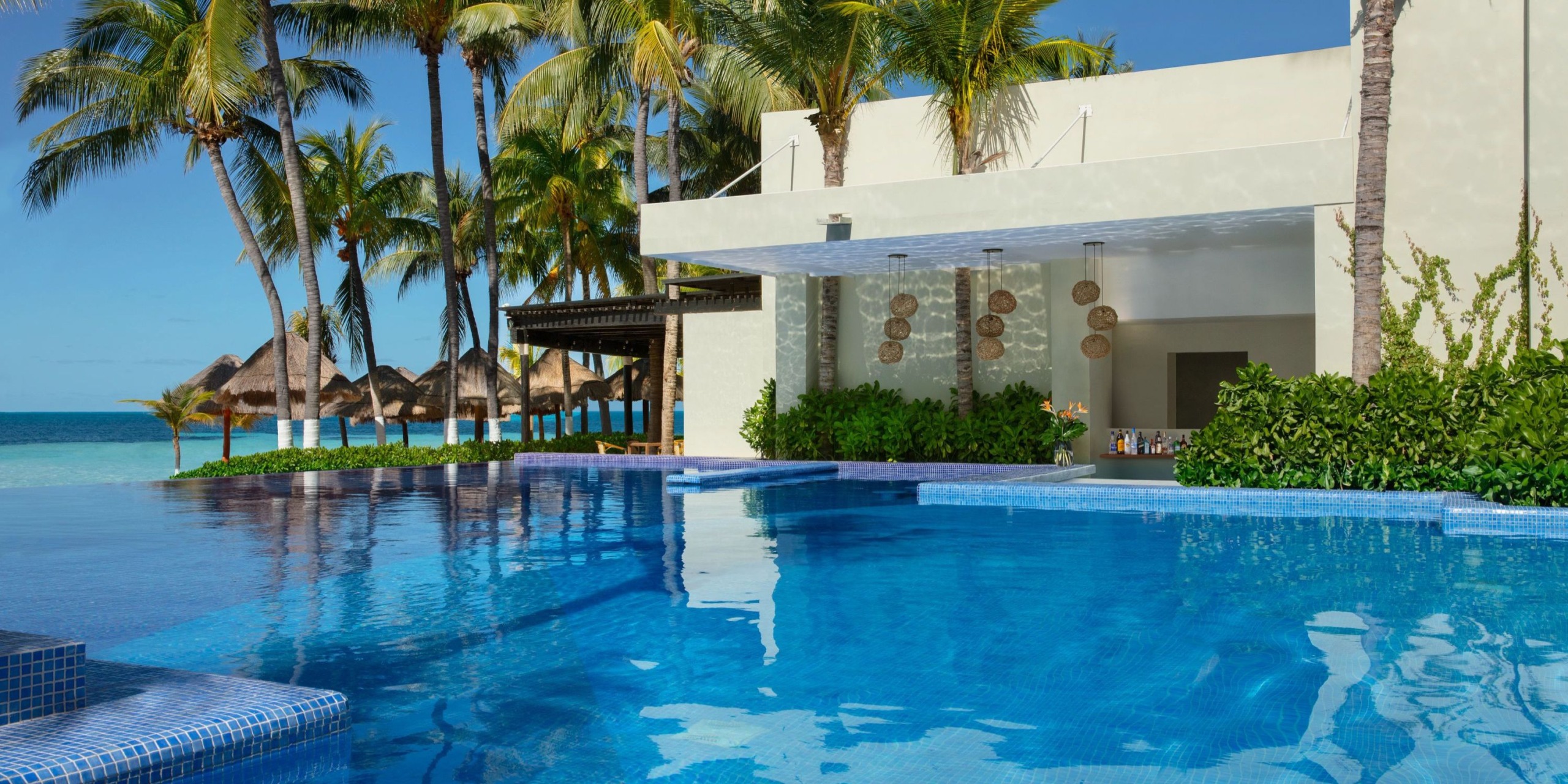 The Dreams Sands Cancun features an infinity pool beside a sleek, modern white structure surrounded by palm trees, offering a view of a beach adorned with straw umbrellas beneath a clear blue sky.