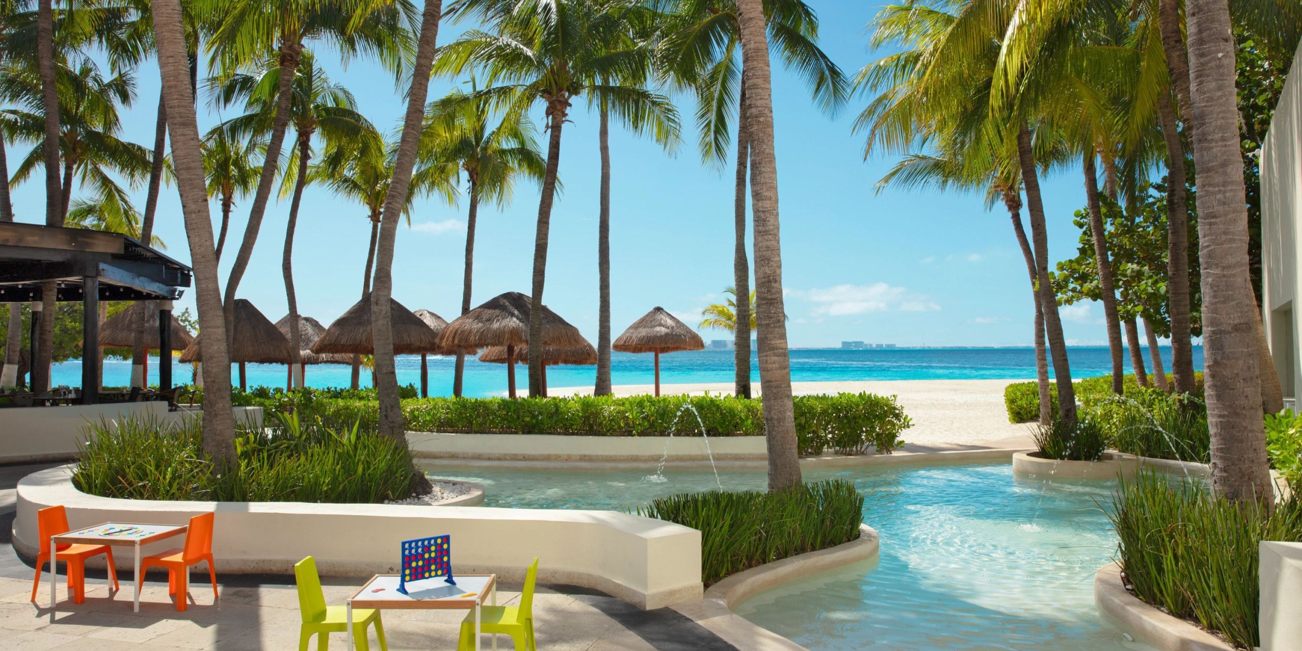 A tropical outdoor scene at Dreams Sands Cancun, featuring palm trees, thatched beach umbrellas, a pool, and colorful patio furniture under a clear blue sky.