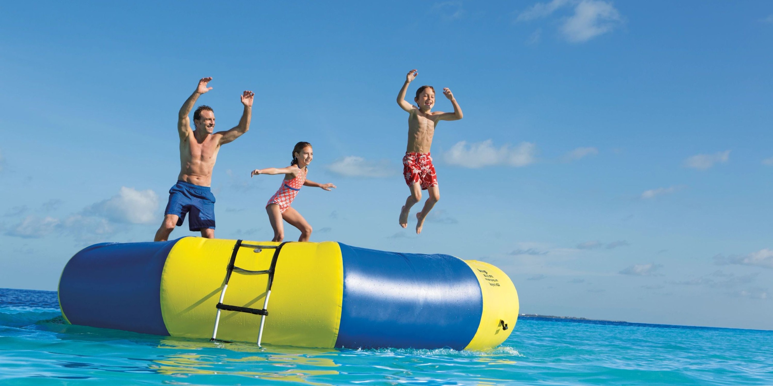 Three people leap from the Dreams Sands Cancun inflatable platform into the ocean on a clear day.