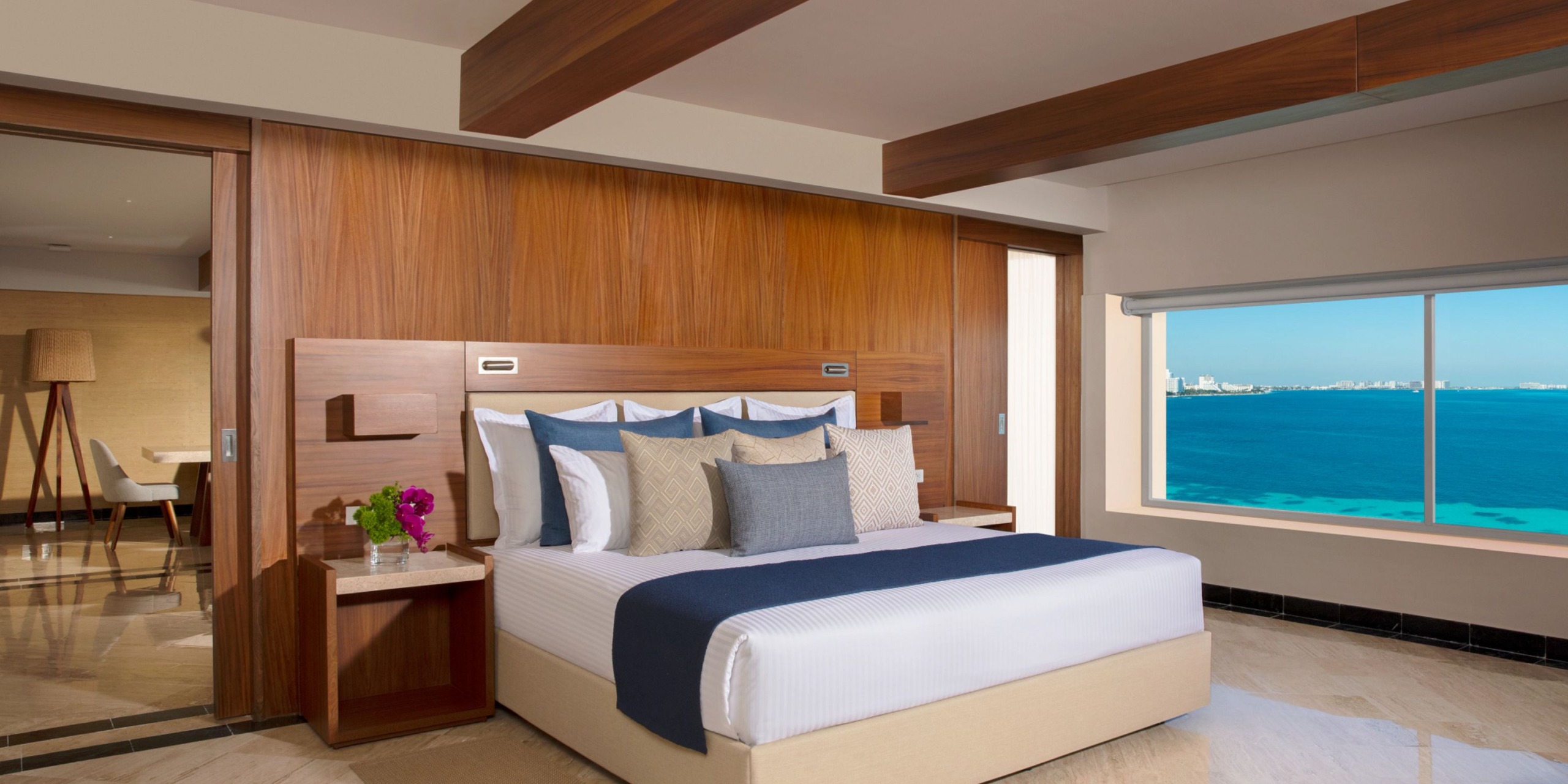 Experience the Dreams Sands Cancun, featuring a hotel room with a large bed adorned with decorative pillows, elegant wooden accents, and a breathtaking open view of the ocean through the window. A charming small table with flowers is thoughtfully placed beside the bed.