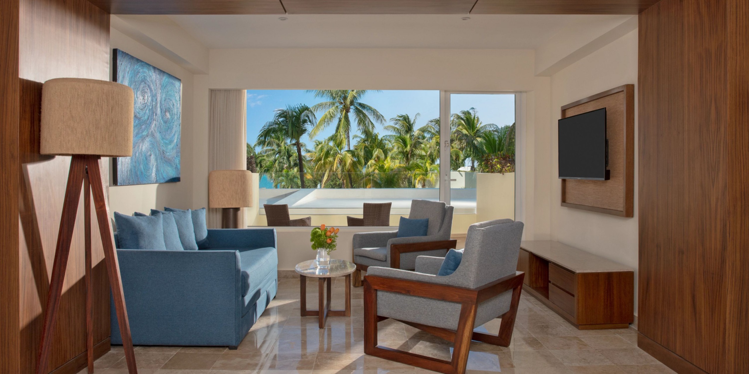 A modern living room at Dreams Sands Cancun features blue sofas, wooden chairs, and a view of palm trees through a large window. A TV is mounted on the wall, and two lamps flank the seating area.