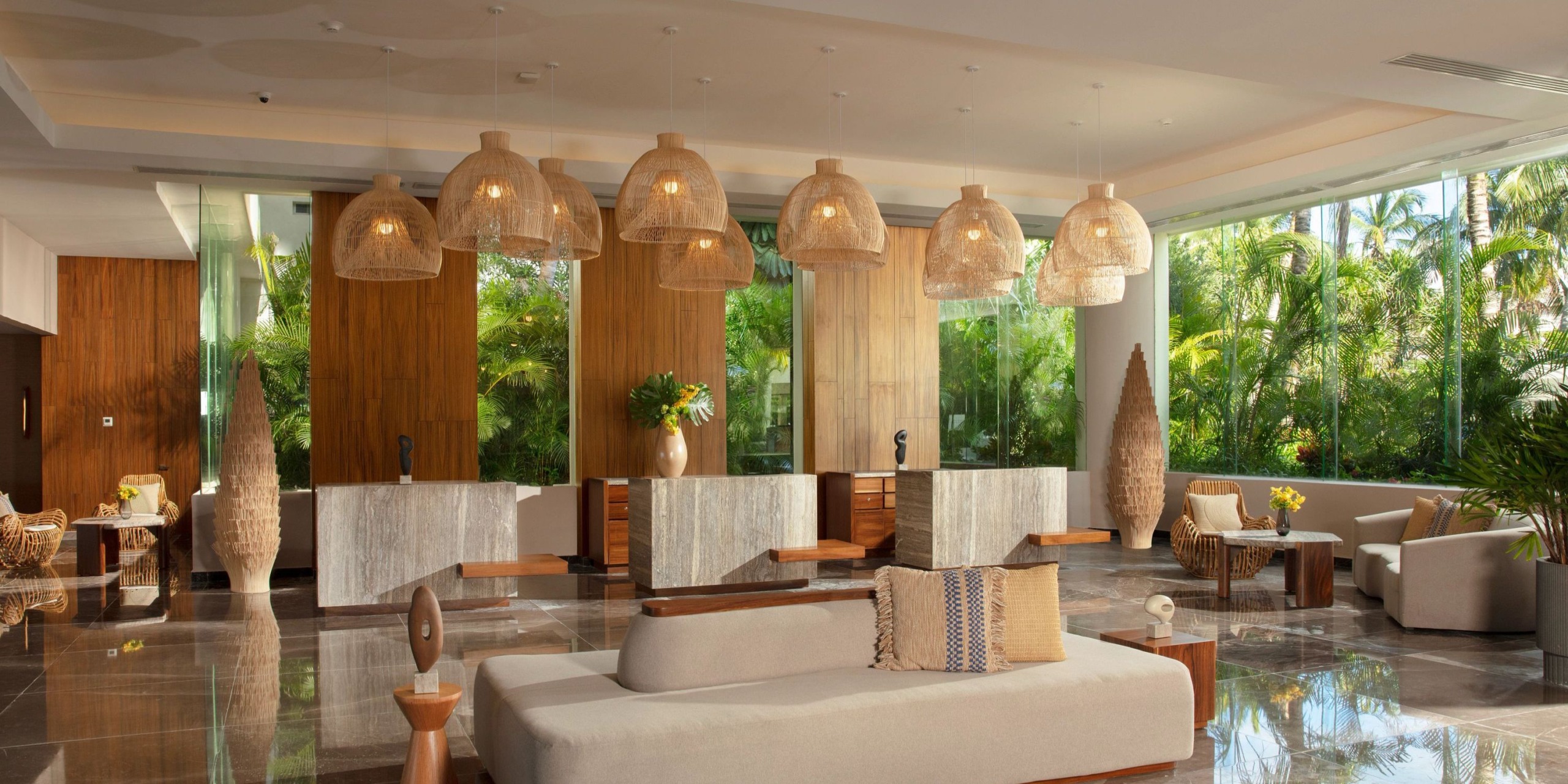 The Dreams Sands Cancun lobby offers a spacious and modern decor, featuring wicker light fixtures, wooden accents, large windows overlooking the garden, and a central seating area adorned with neutral-colored furniture.